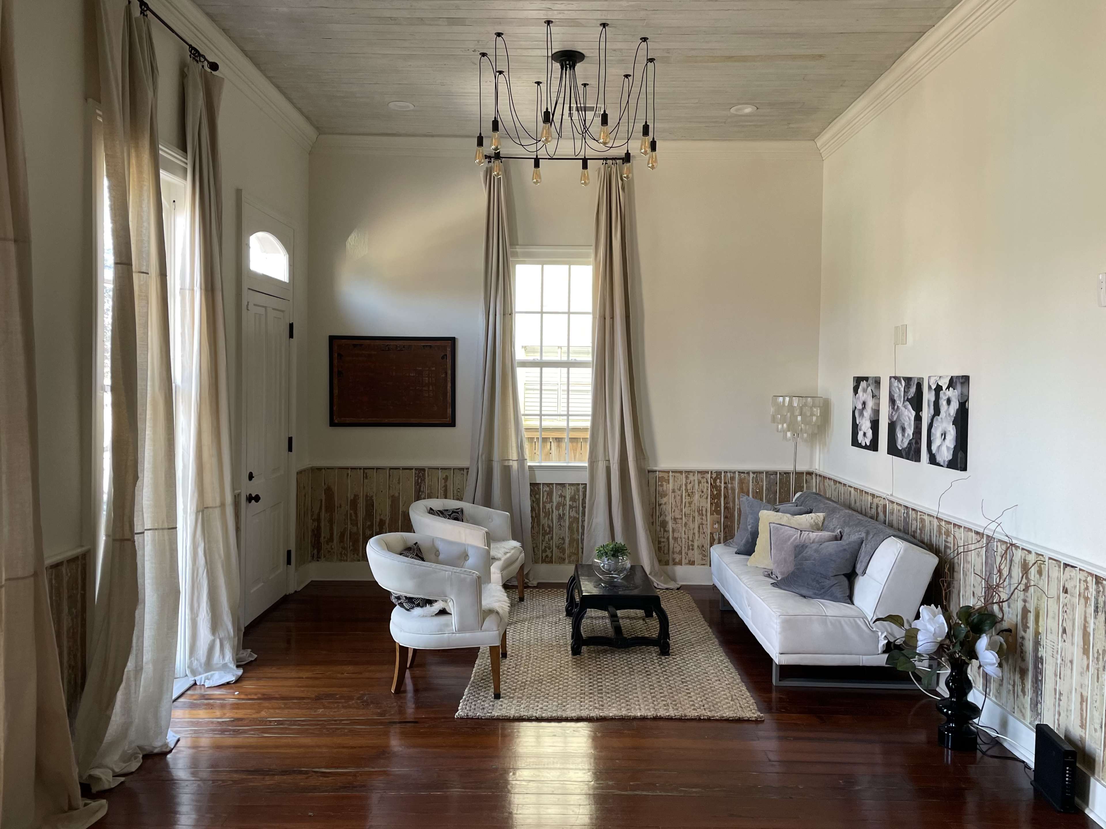 A bright living room with a pair of white armchairs and a sofa, accompanied by a coffee table on a woven rug, surrounded by tall curtains and wooden flooring.