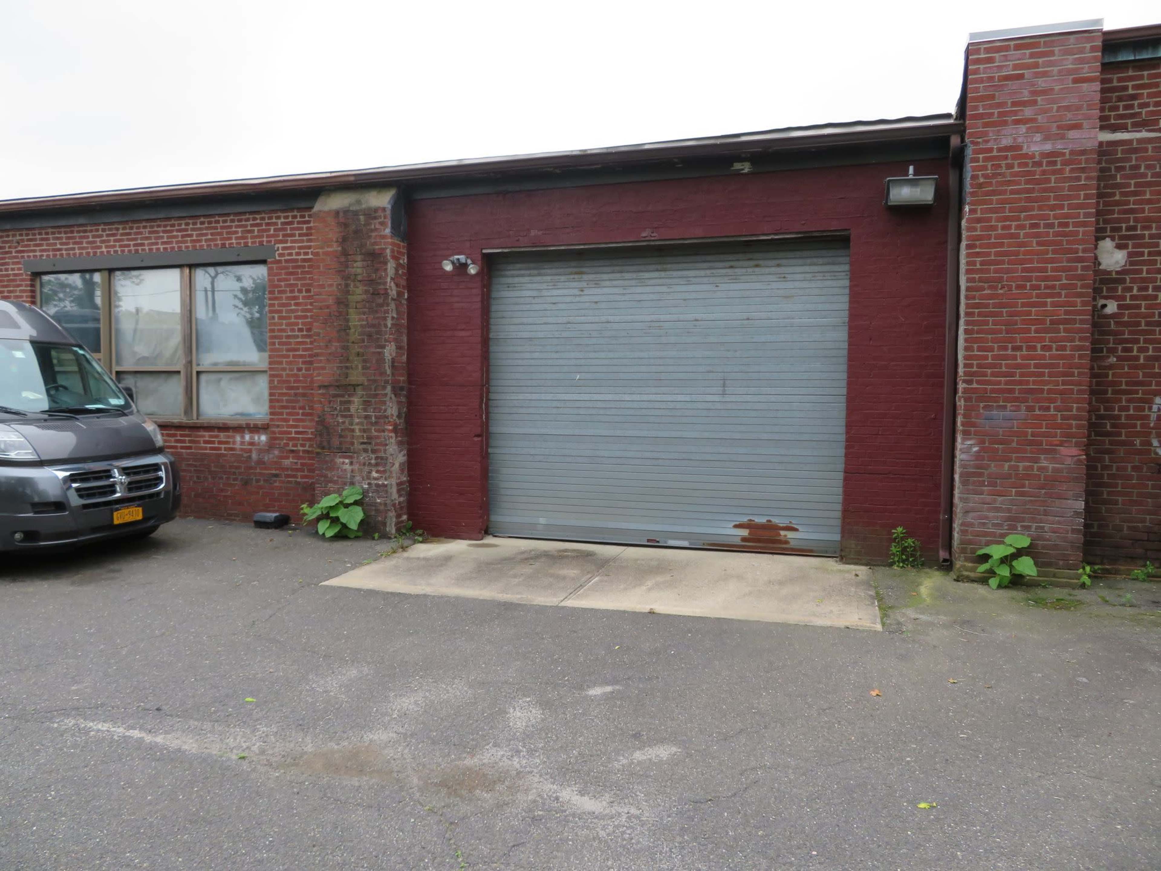 The image shows a red brick building with a gray garage door and a van parked nearby, surrounded by a concrete area with some greenery.