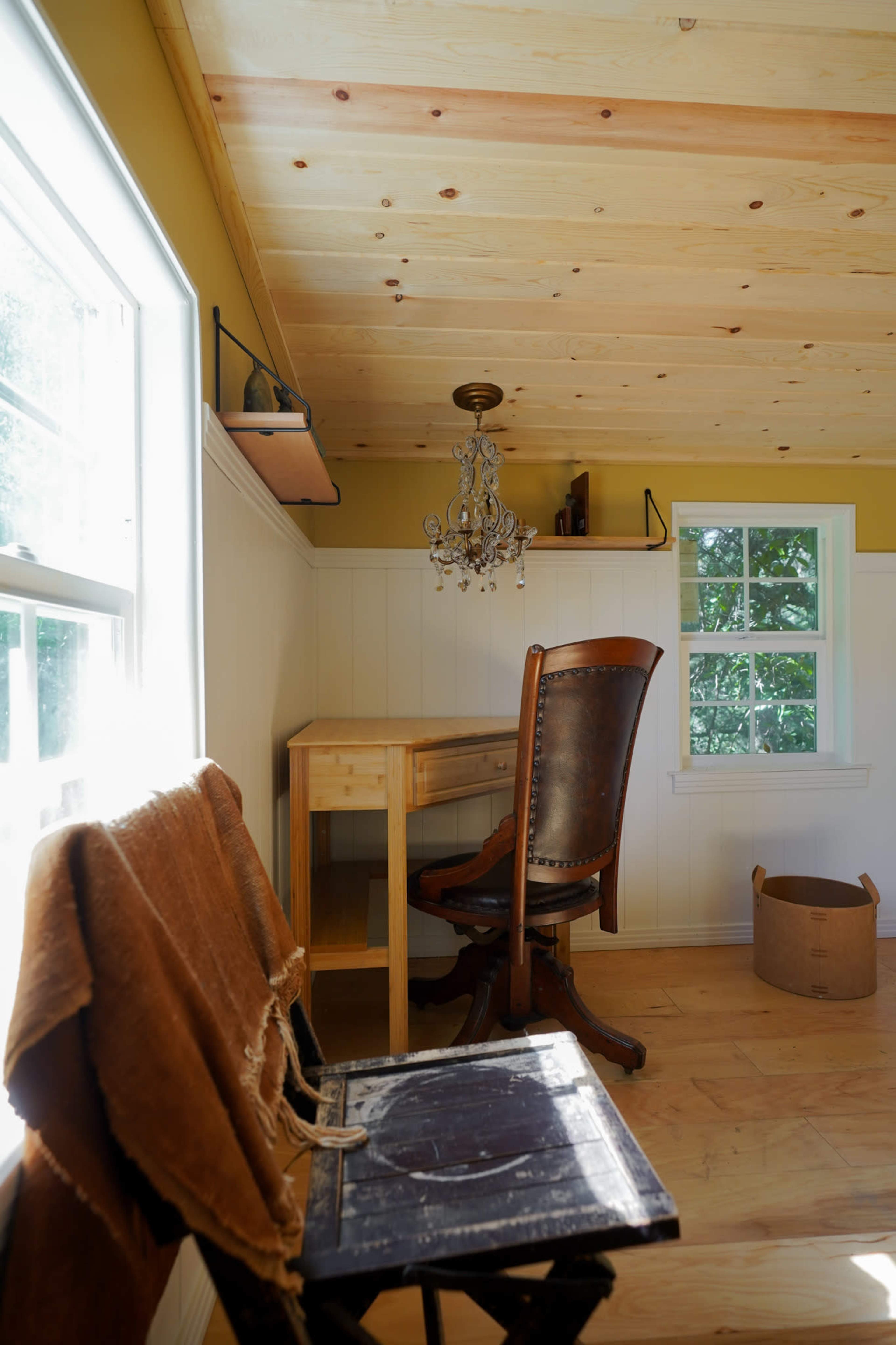 The image shows a well-lit, simple room featuring a wooden desk and a leather chair, with a chandelier suspended from the ceiling and a window providing natural light.