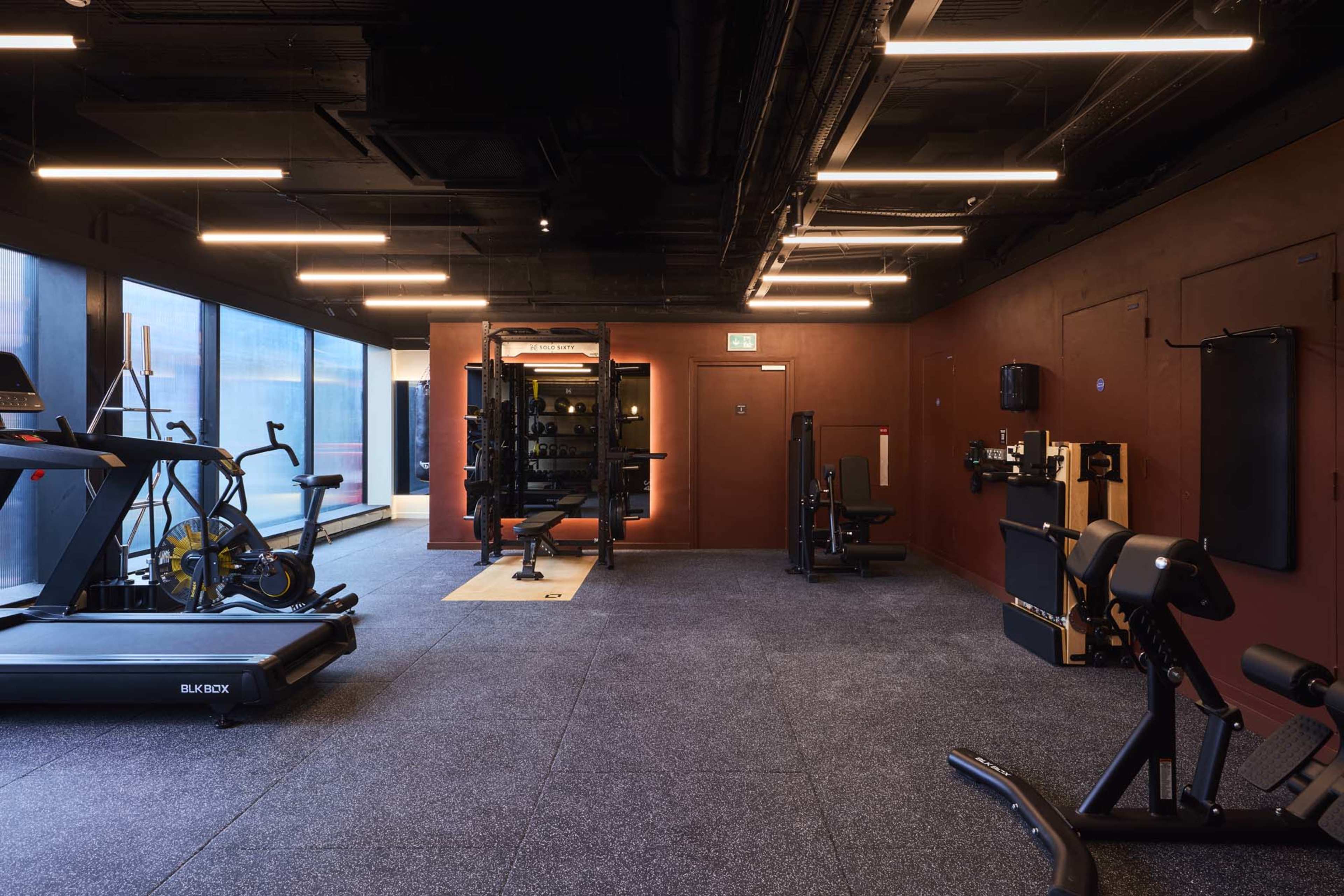 The image shows a modern gym interior with various exercise equipment, including a treadmill, weight machine, and strength training benches on a rubber floor.