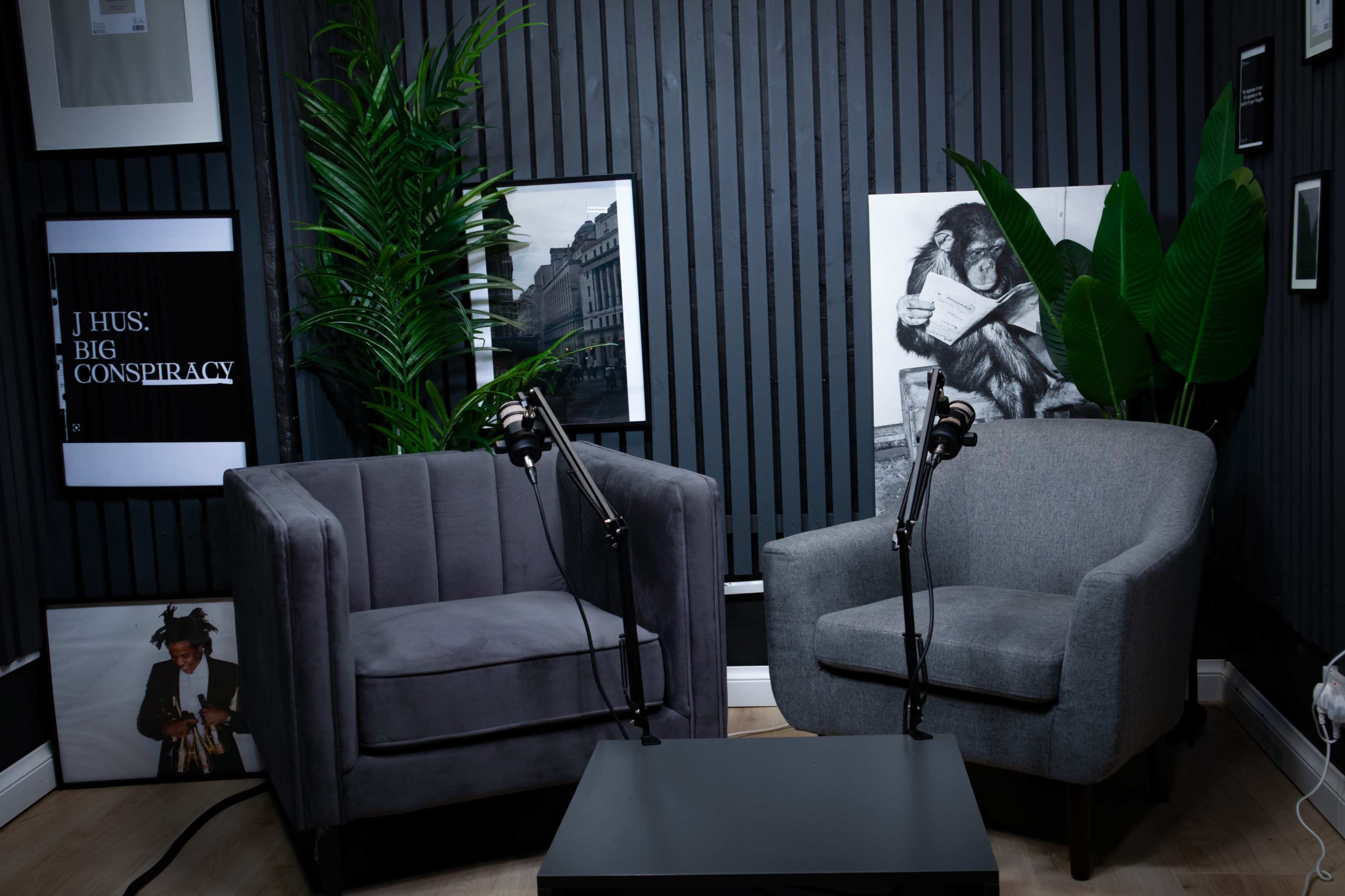 The image shows a podcast recording setup featuring two armchairs with microphones on stands, surrounded by framed artwork and greenery on a striped wall.