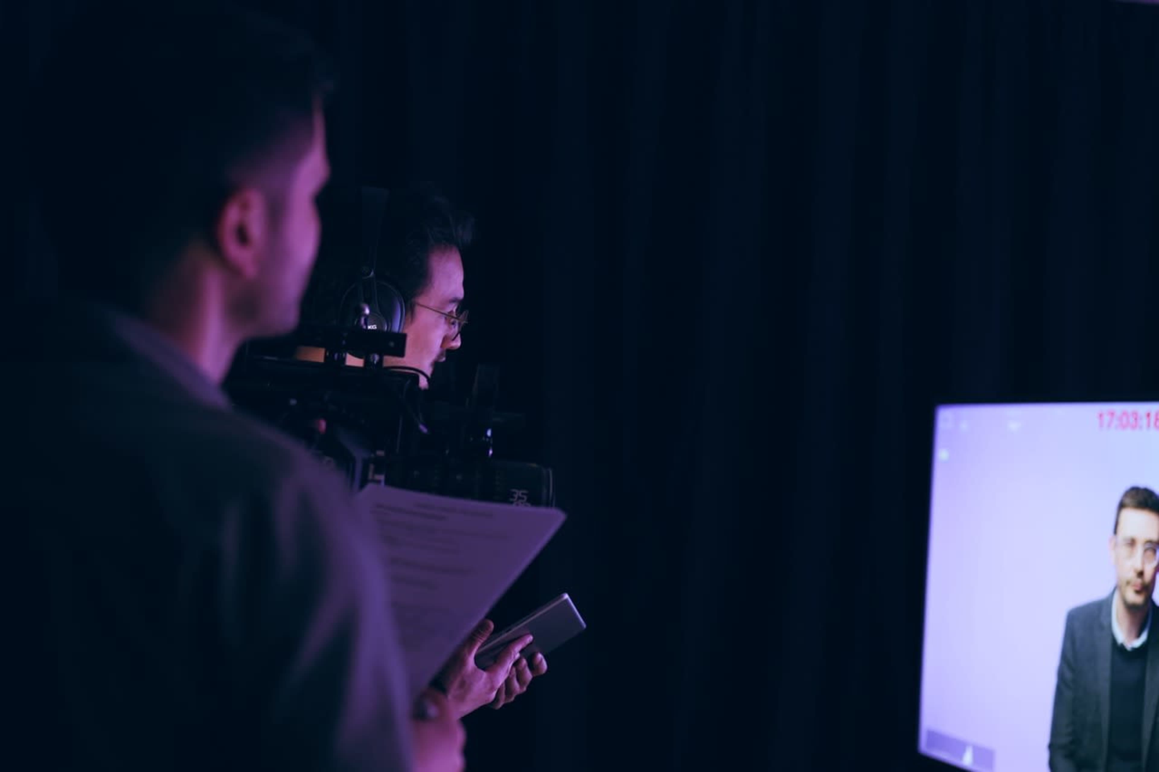 A person holding a script stands in front of a monitor displaying another individual during a video shoot.