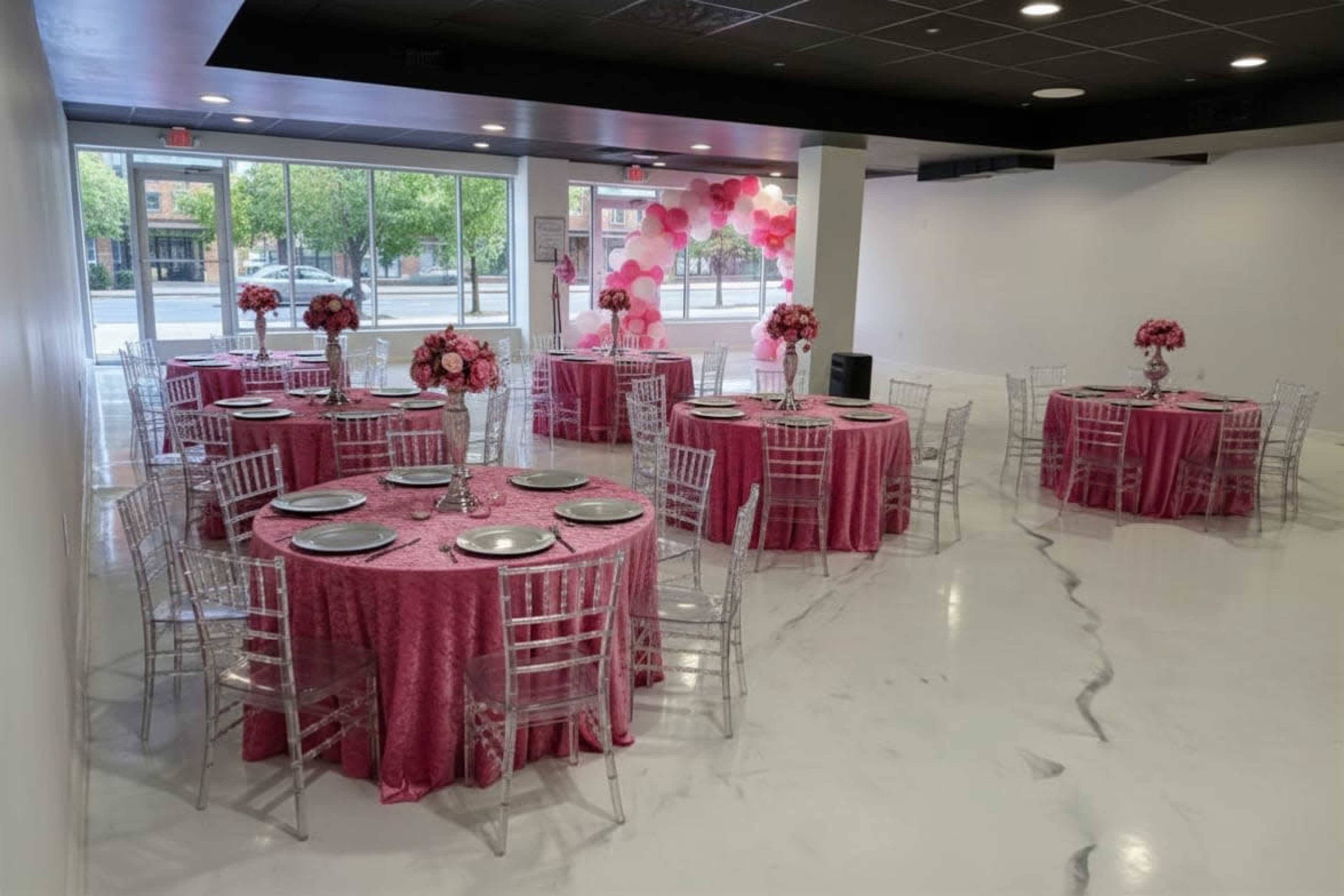 The image shows a banquet hall arranged with several round tables covered in pink tablecloths, each adorned with floral centerpieces and surrounded by clear acrylic chairs.