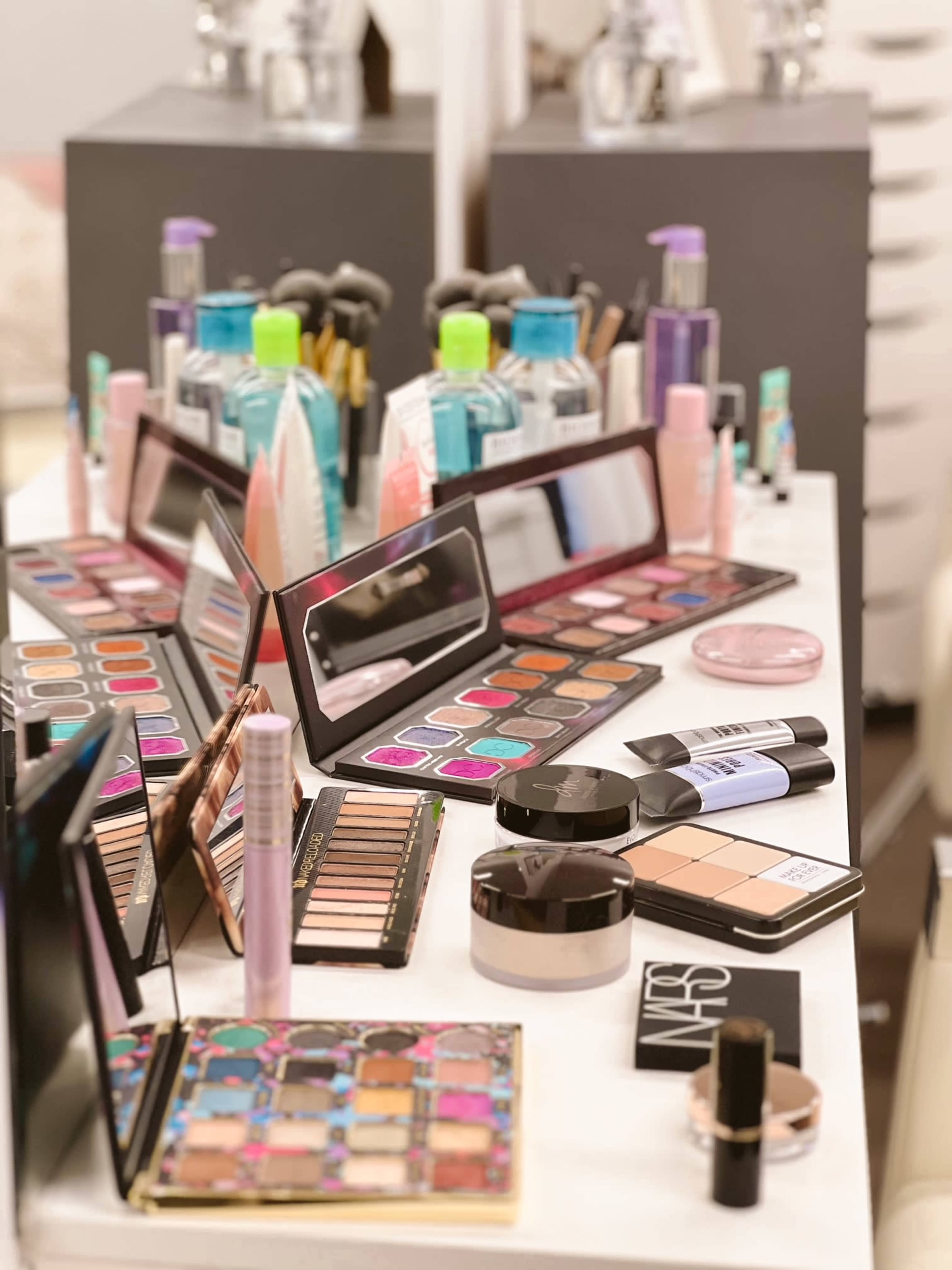 A cluttered makeup table filled with various palettes, bottles, and beauty products.