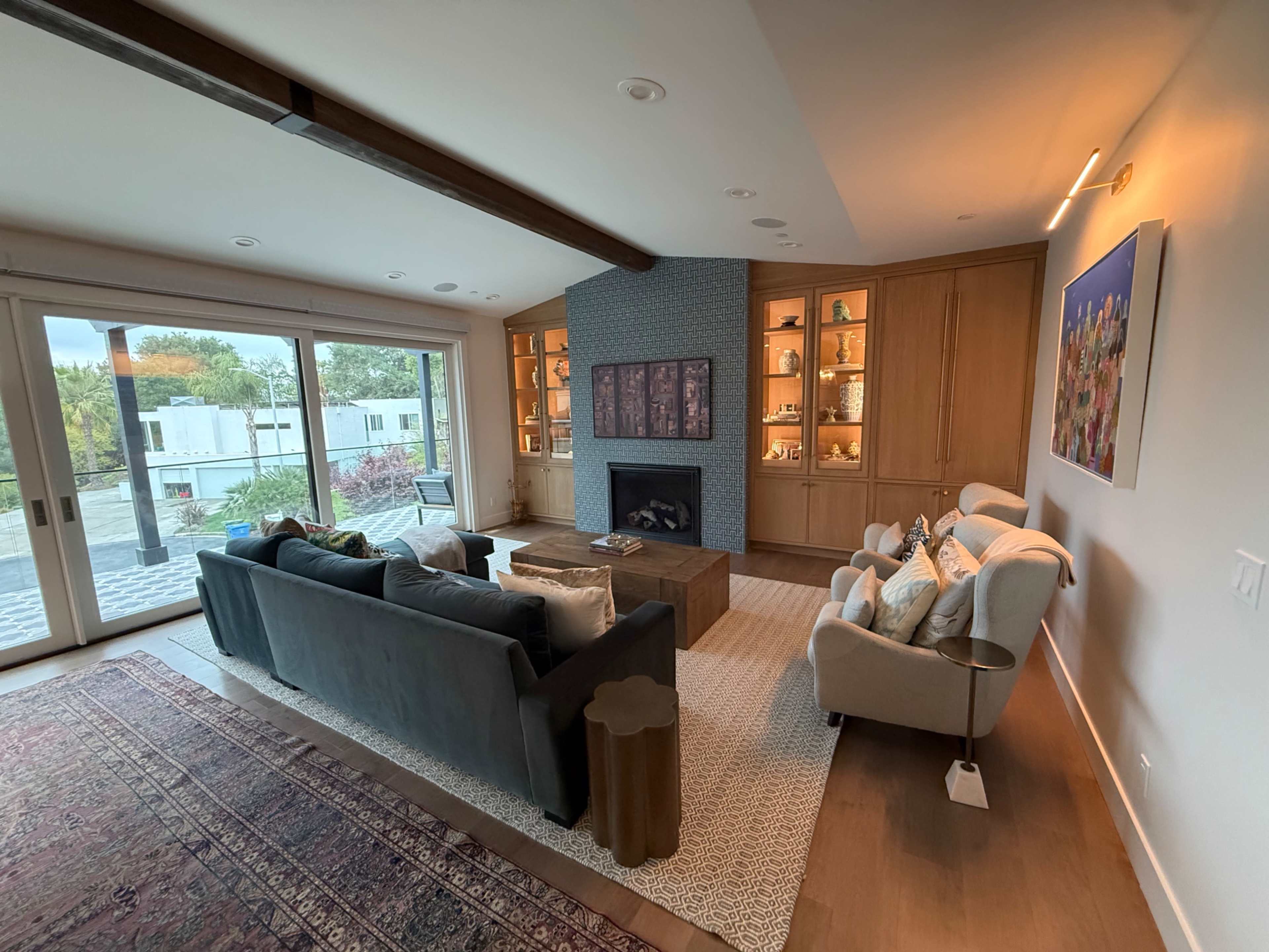 The image shows a modern living room featuring a blue couch, two armchairs, a coffee table, a fireplace with built-in shelving, and large sliding glass doors leading to an outdoor space.