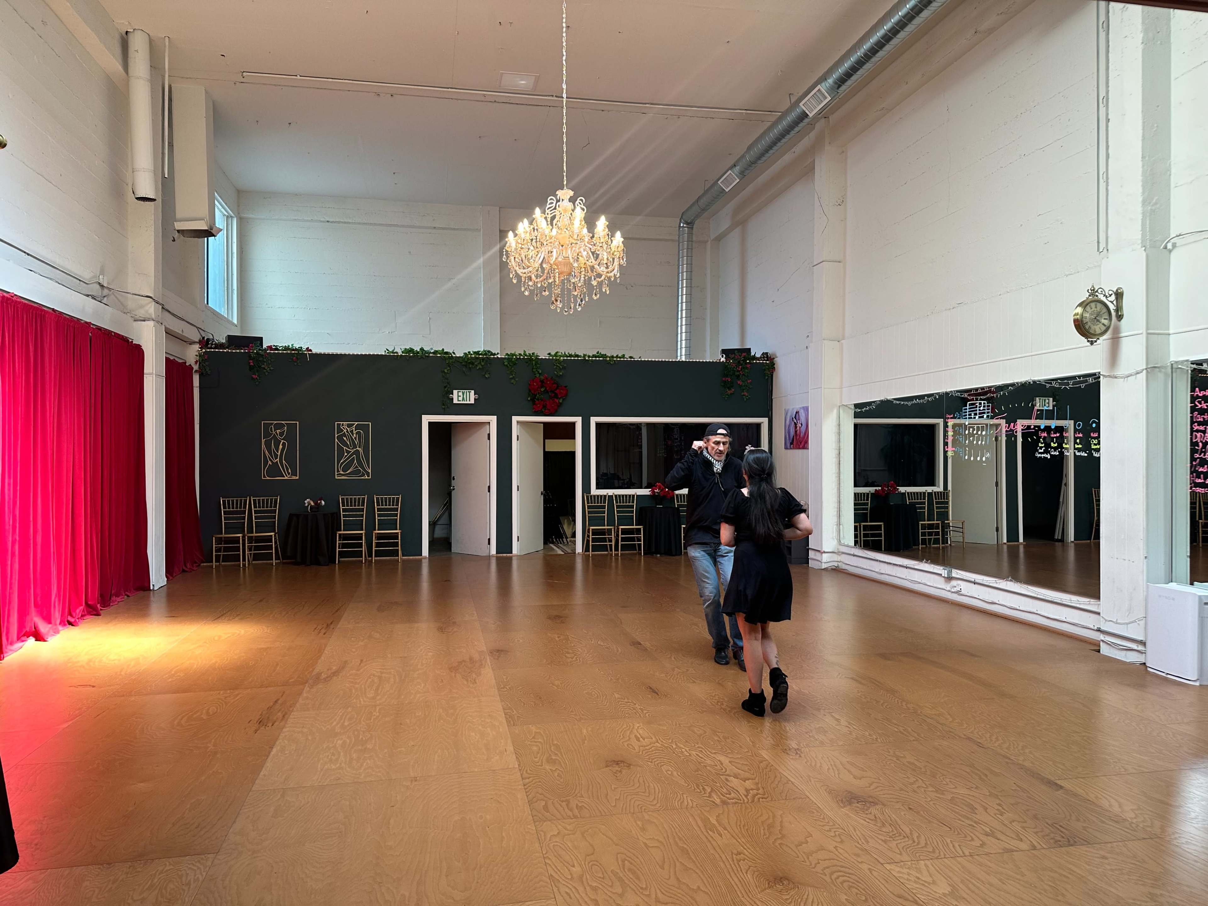 A spacious dance studio features a wooden floor, large mirrors, and a chandelier, with two people dancing in the center.