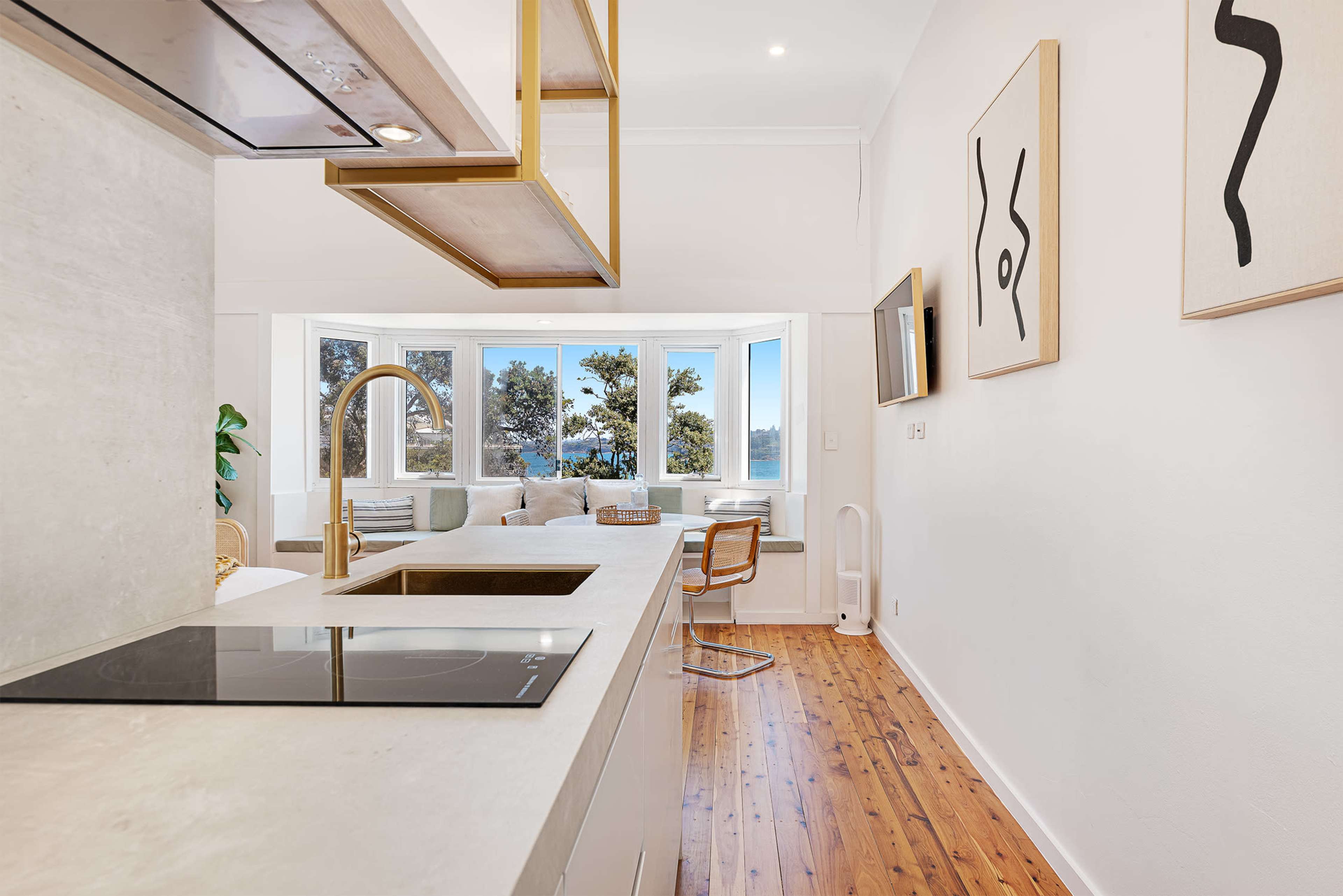 A modern kitchen that opens into a living area with large windows overlooking a view, featuring wooden flooring and minimalistic decor.
