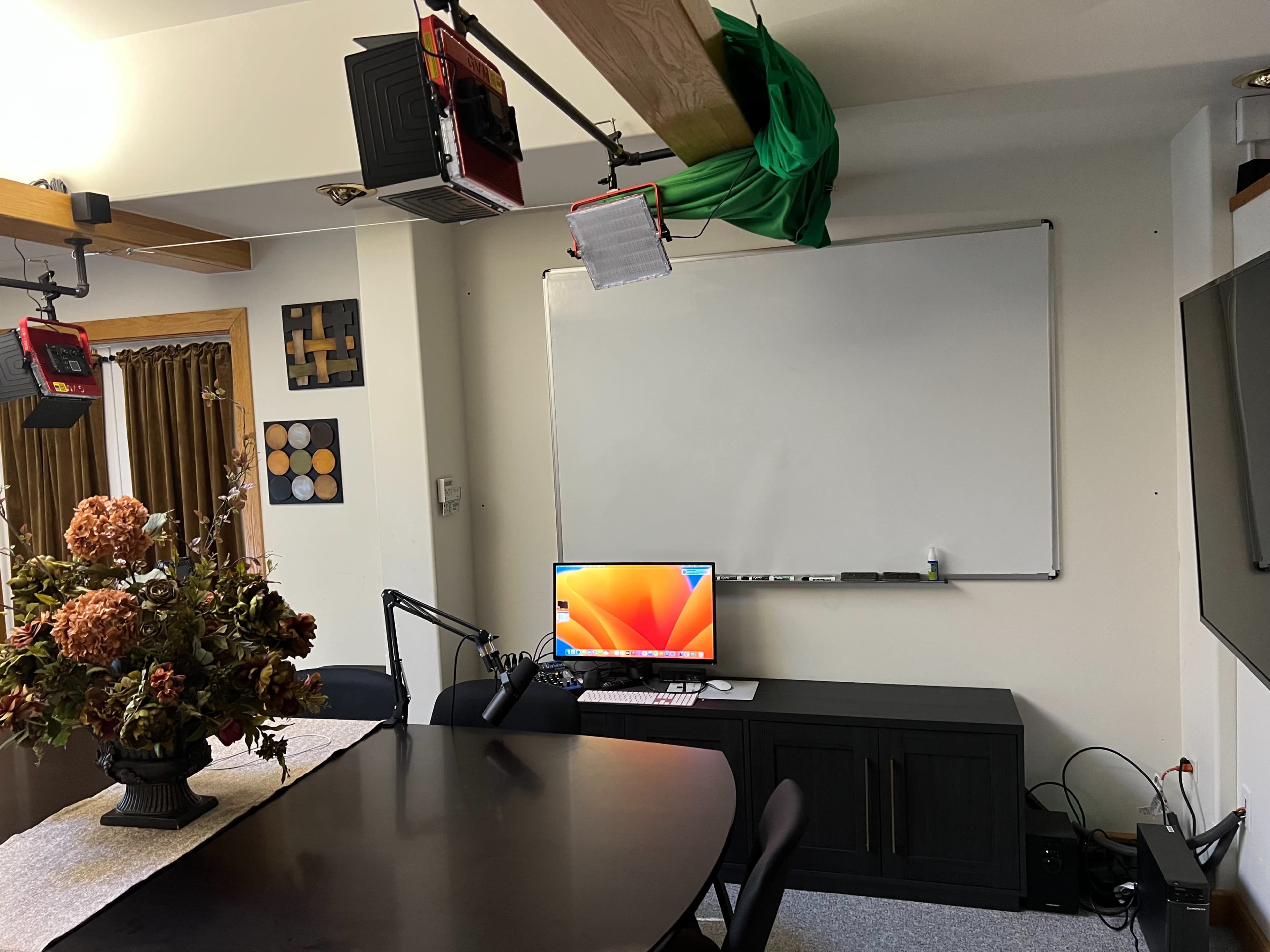 A room with a large table, a monitor on a desk, a whiteboard mounted on the wall, and studio lights hanging from the ceiling.