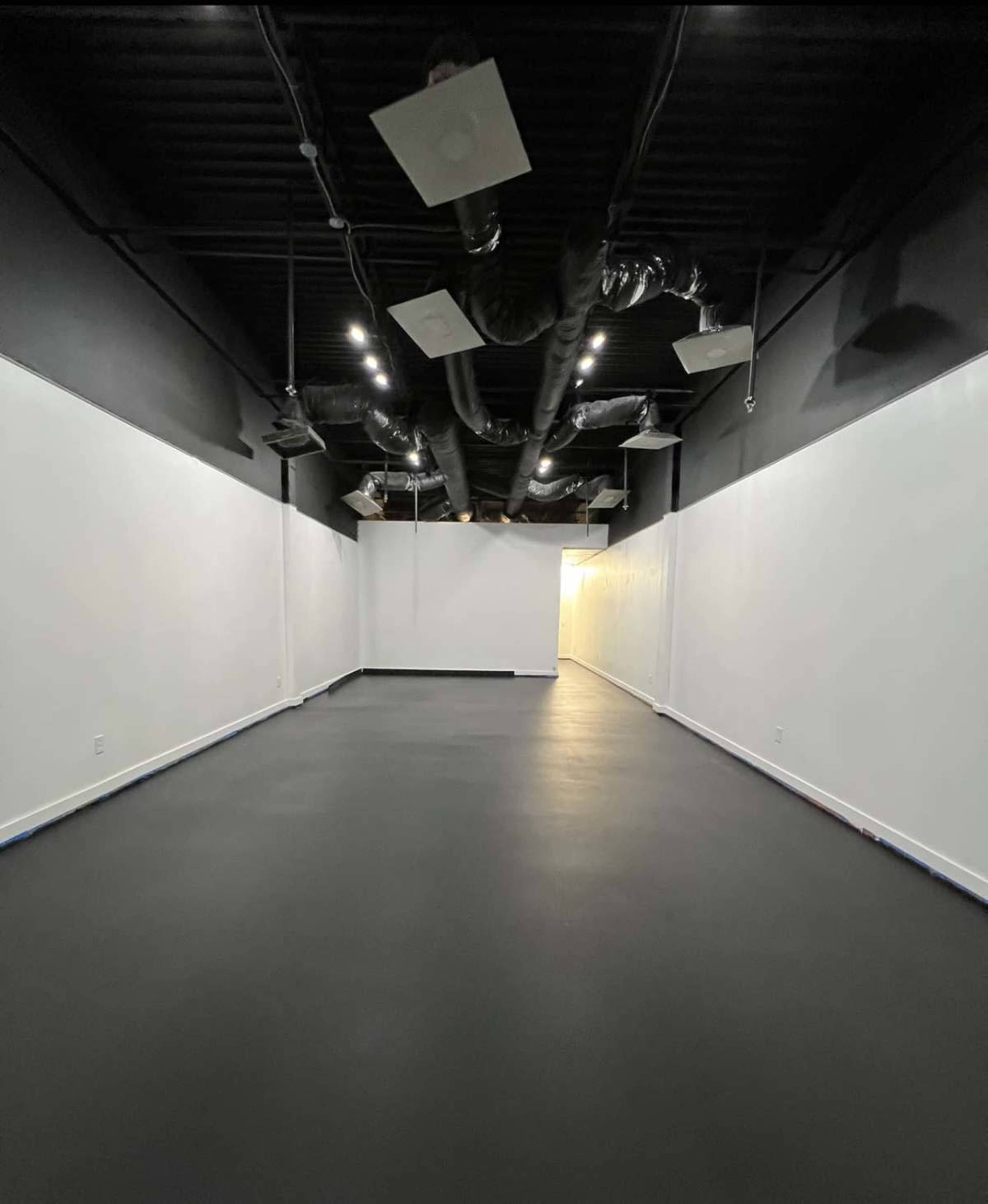 The image shows a spacious, empty room with black walls and a smooth black floor, featuring ductwork and lights mounted on the ceiling.