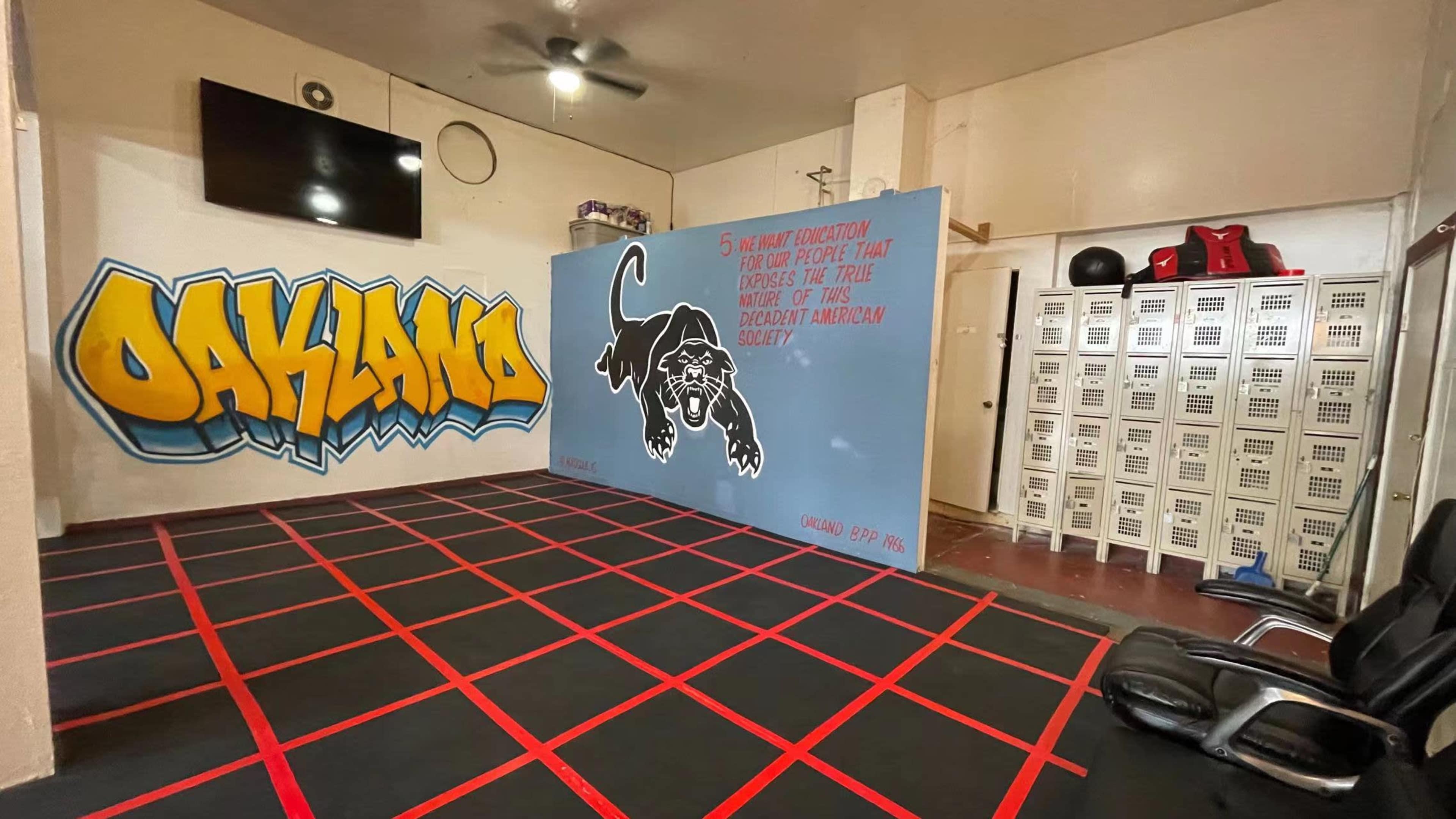 A gym interior with a graffiti mural reading "OAKLAND" on one wall, a quote on another wall featuring a black panther, and a row of lockers in the background.