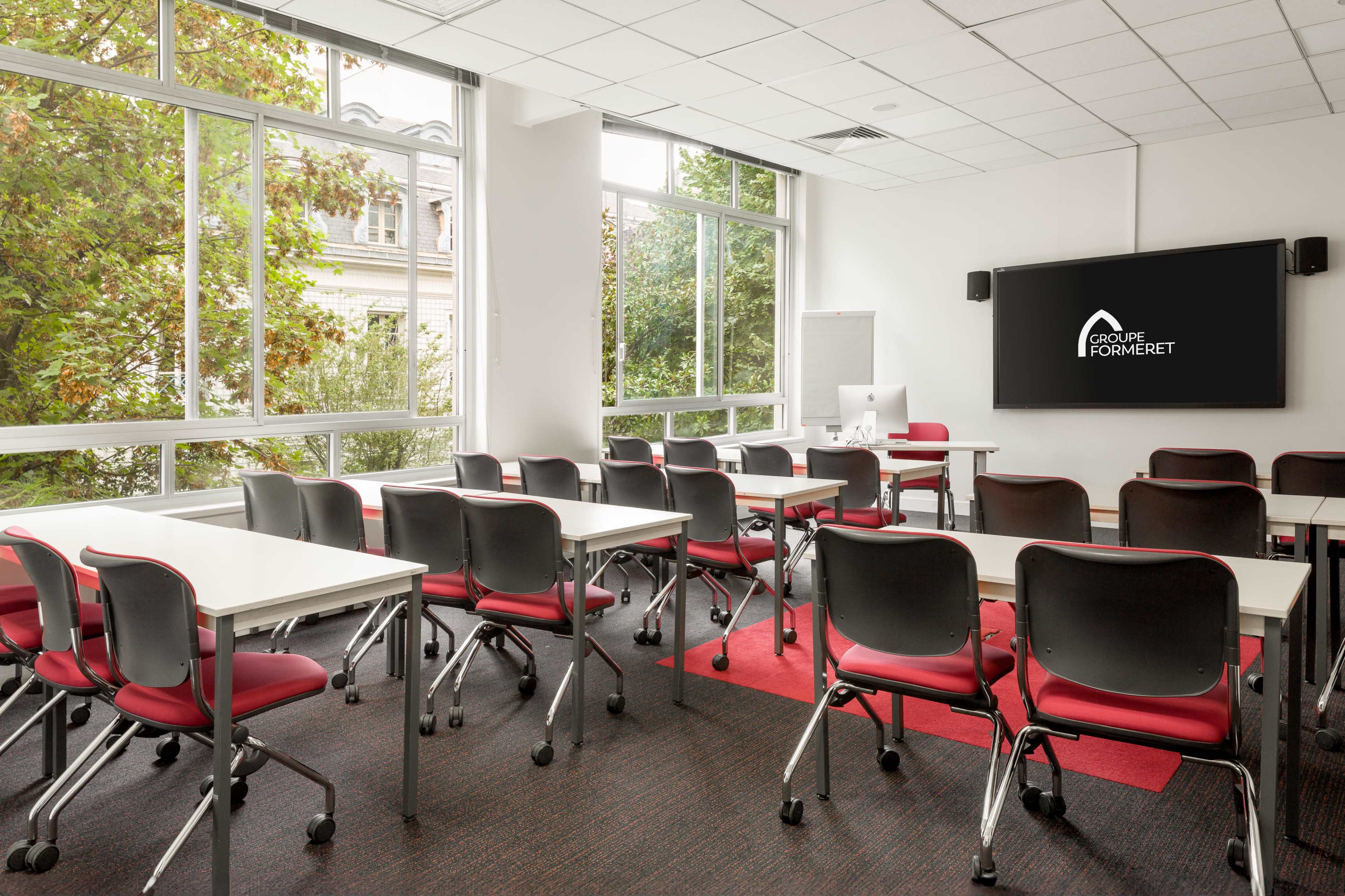 A modern classroom is arranged with rows of red and black chairs and white tables, featuring large windows that provide natural light and a wall-mounted screen displaying a logo.