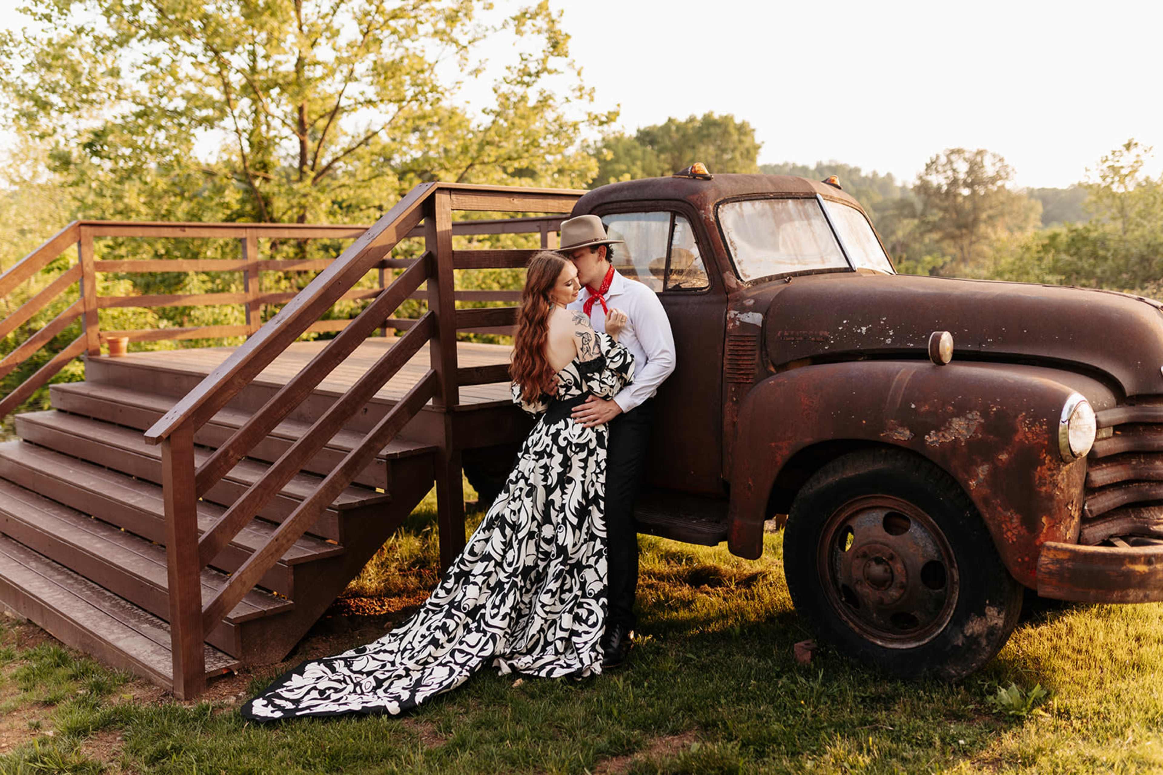 A couple poses near an old, rusty pickup truck next to a wooden staircase in a lush green outdoor setting.