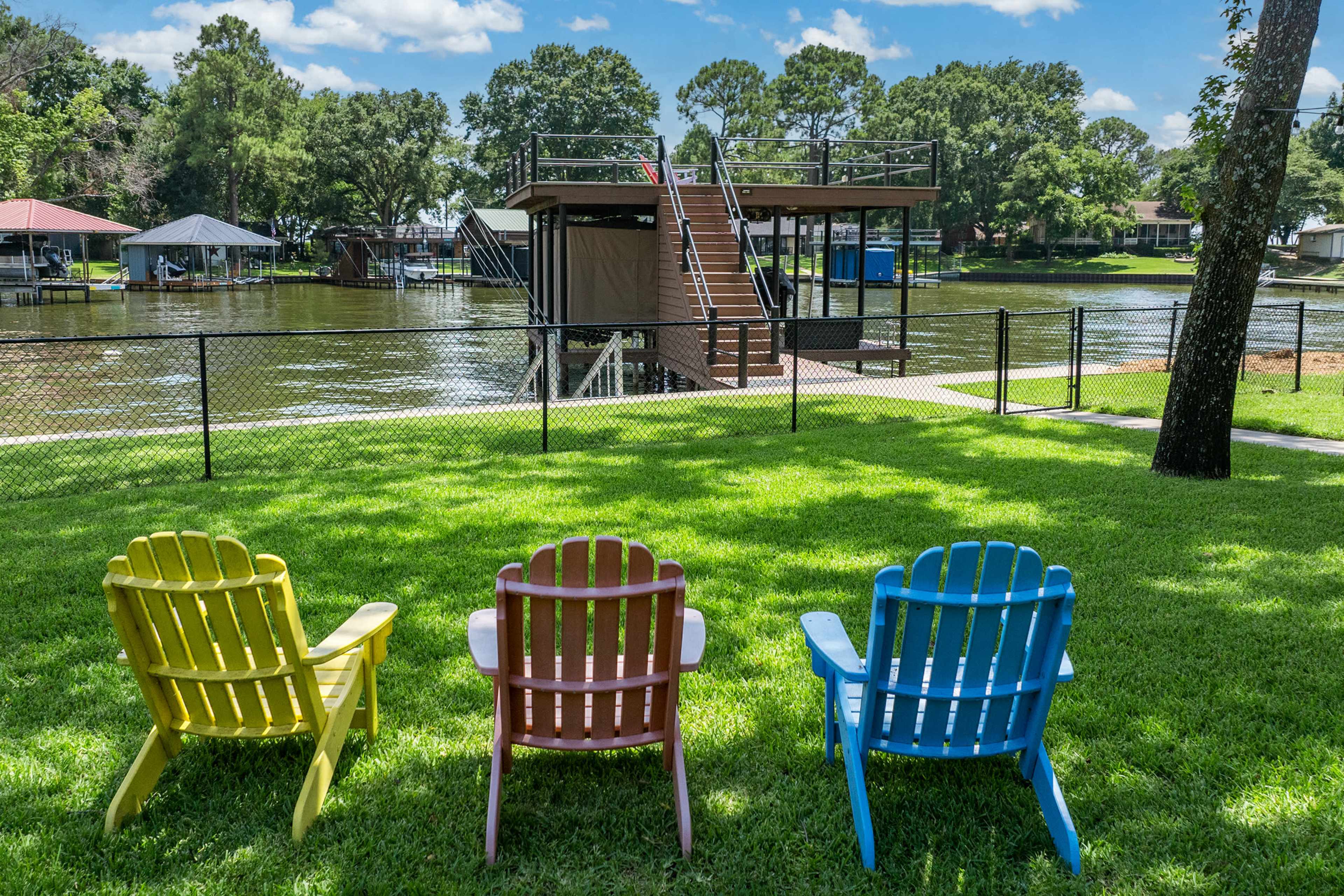 Three colorful Adirondack chairs sit on green grass facing a lake, with a dock and stairs visible in the background.