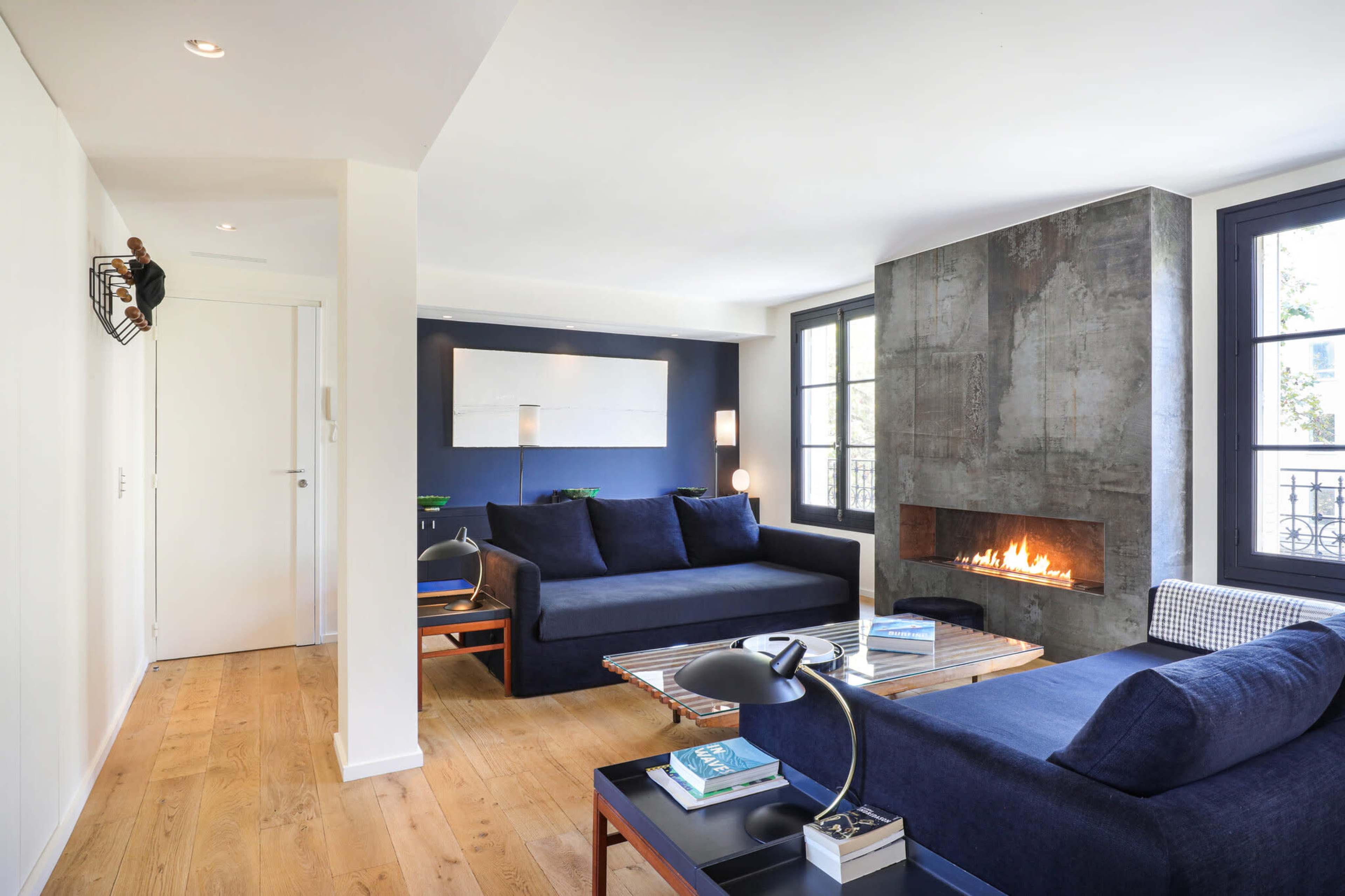 The image shows a modern living room featuring a concrete fireplace, navy blue sofas, and wooden flooring with large windows.