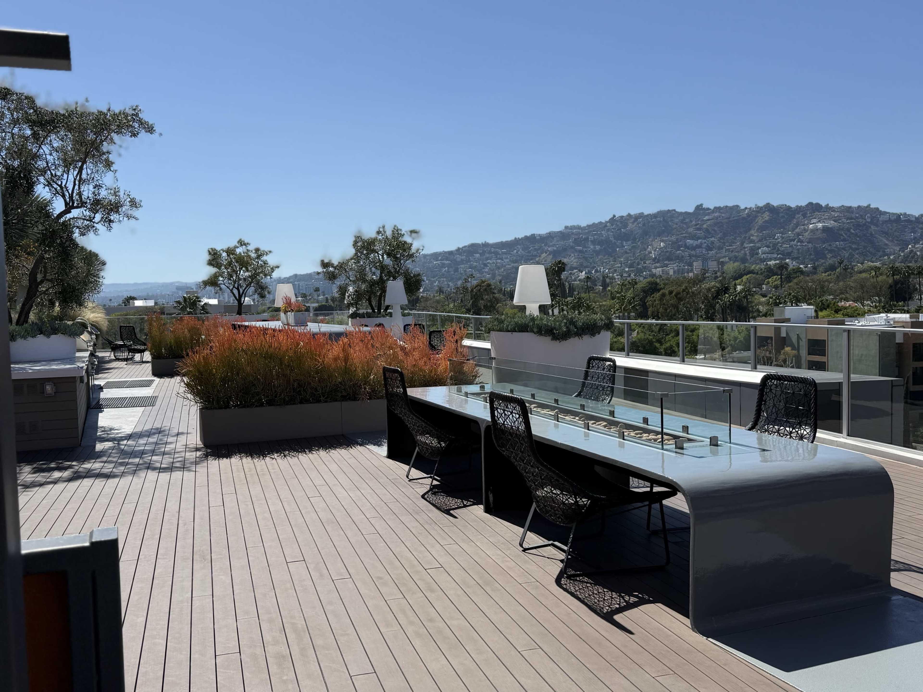 A rooftop terrace features a long table with chairs and colorful landscaping, bordered by a view of hills in the distance under a clear blue sky.