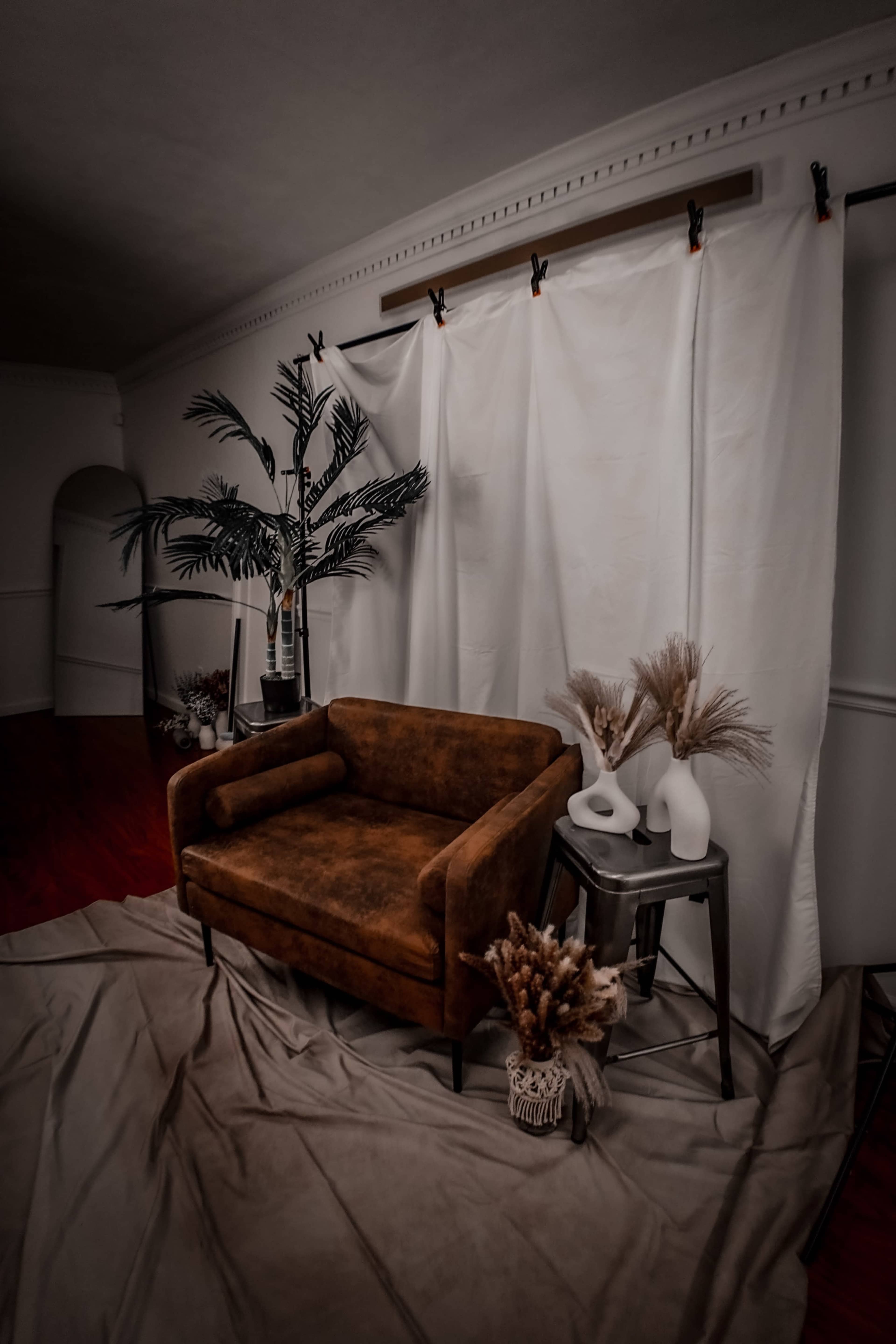A brown couch is placed in front of a white backdrop, accompanied by decorative vases and dried plants on a small table.
