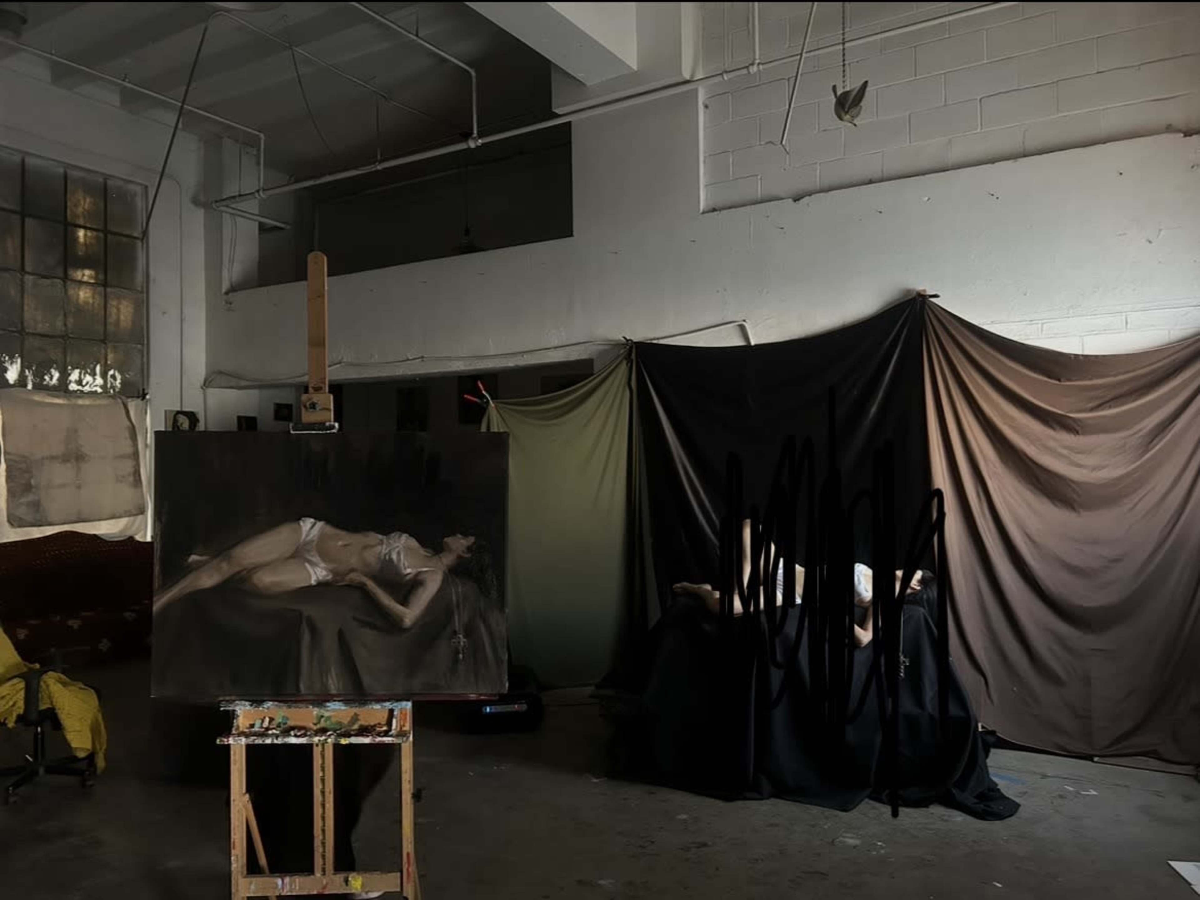 The image depicts an art studio with a partially completed painting of a reclining figure on an easel, while several draped fabrics create a backdrop on one side.