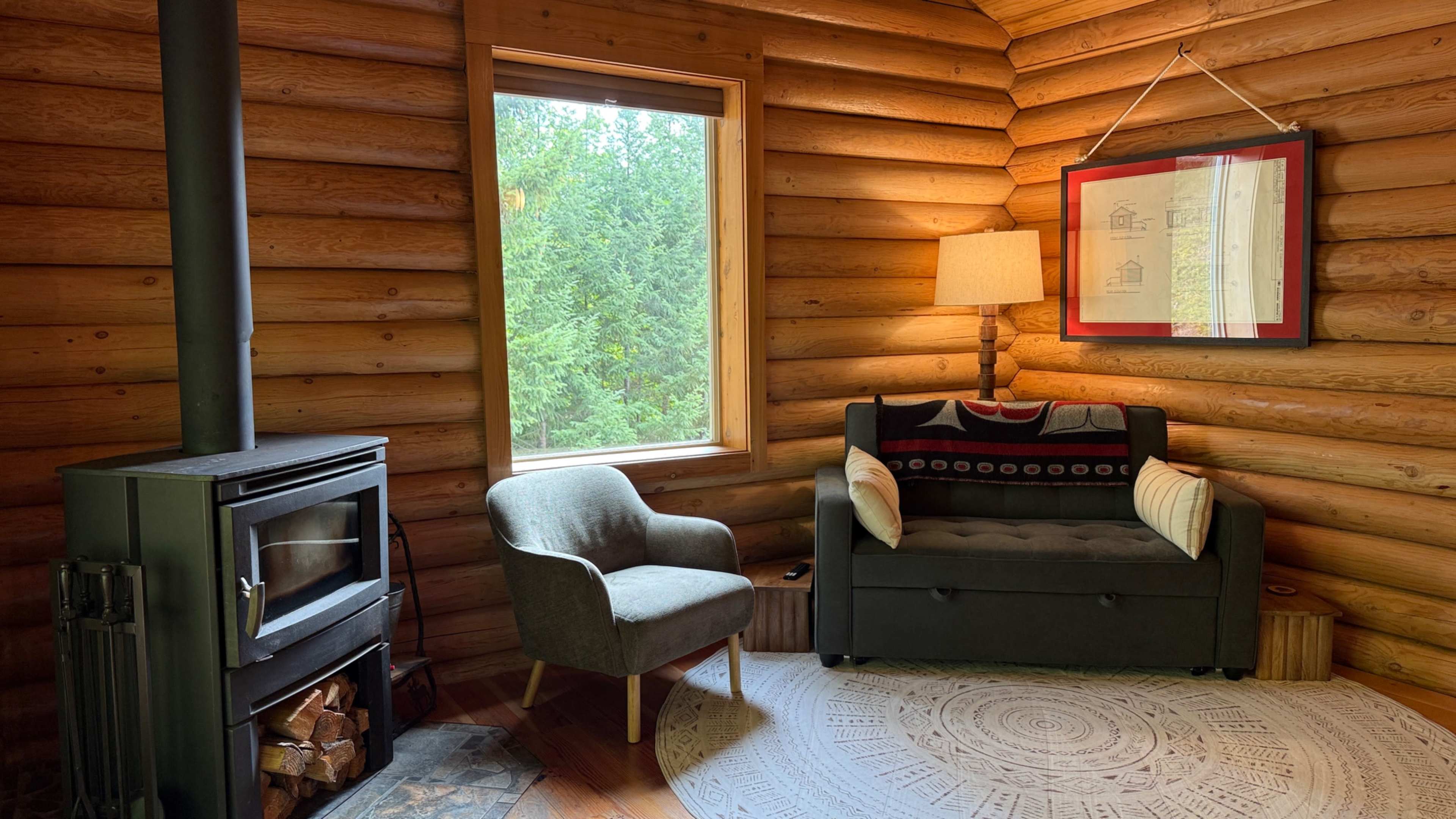 The scene features a cozy corner of a log cabin with a wood-burning stove, a gray armchair, a dark sofa adorned with cushions, and a large window offering a view of green trees outside.