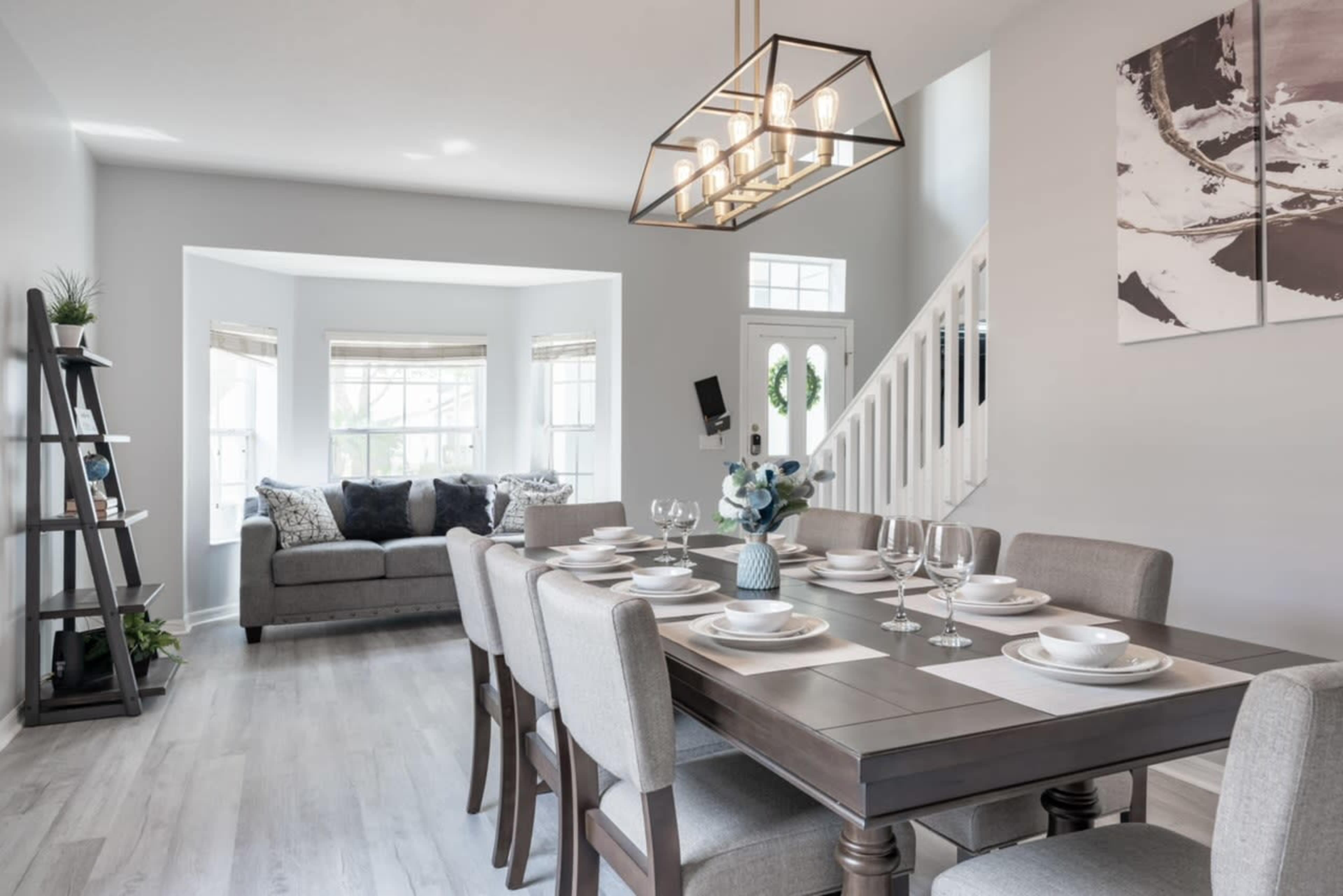 The image shows a modern dining area with a large table set for dinner, surrounded by a light-colored sofa and natural light coming from the windows.