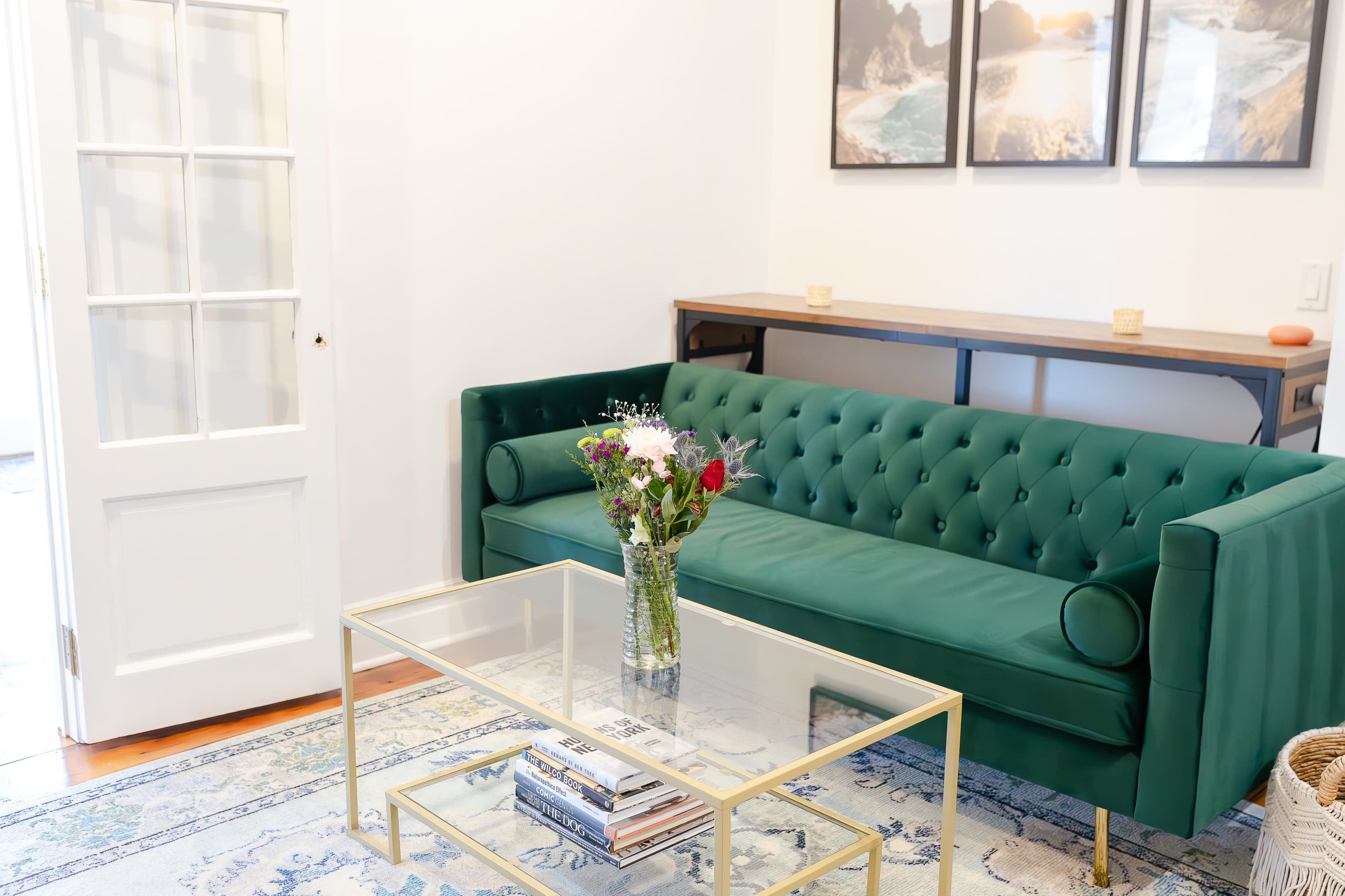 A green tufted sofa sits in a brightly lit room beside a wooden table and a glass coffee table with books and a vase of flowers.