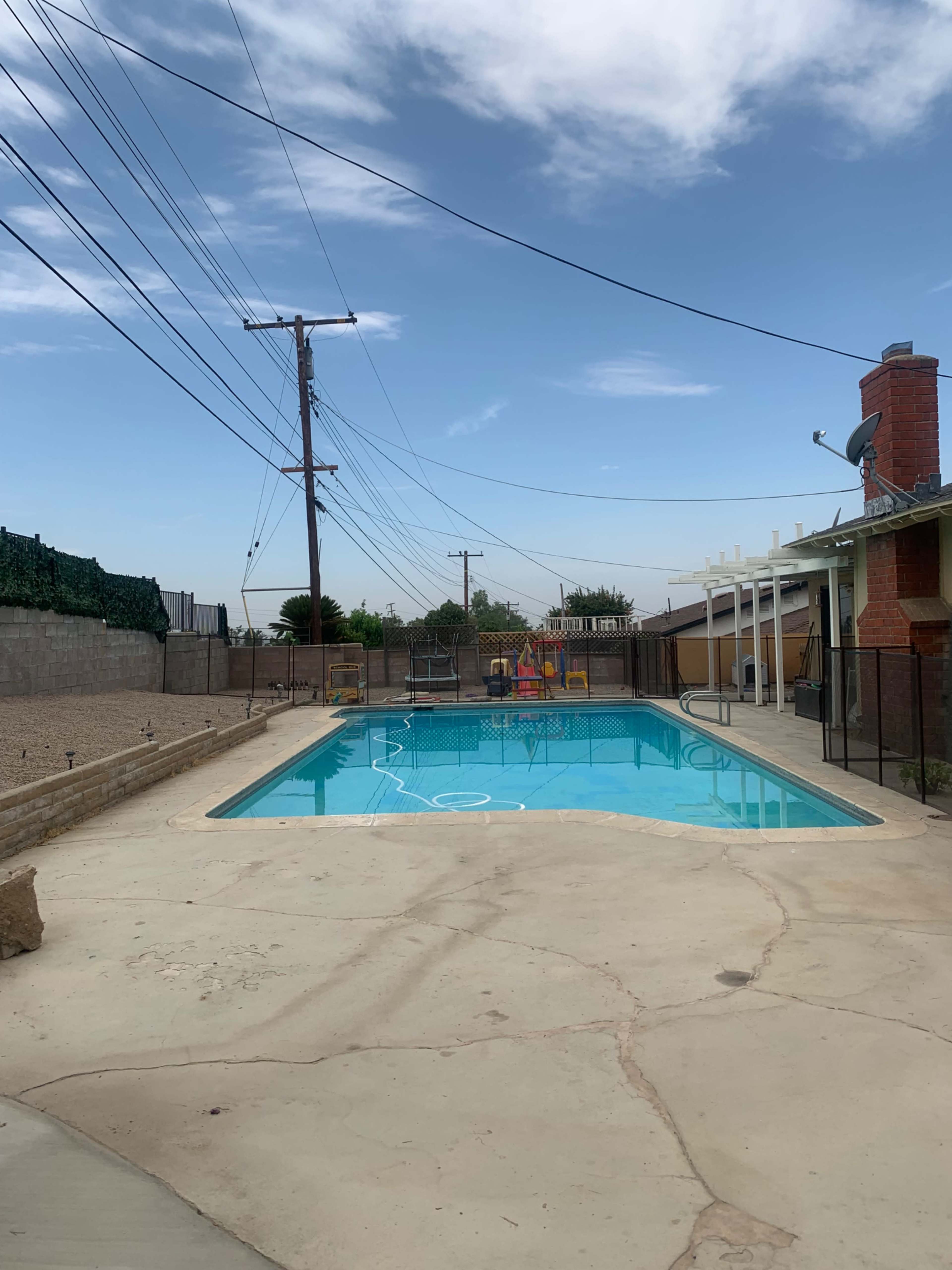 A swimming pool surrounded by a concrete deck and a fence, with power lines overhead and a house in the background.