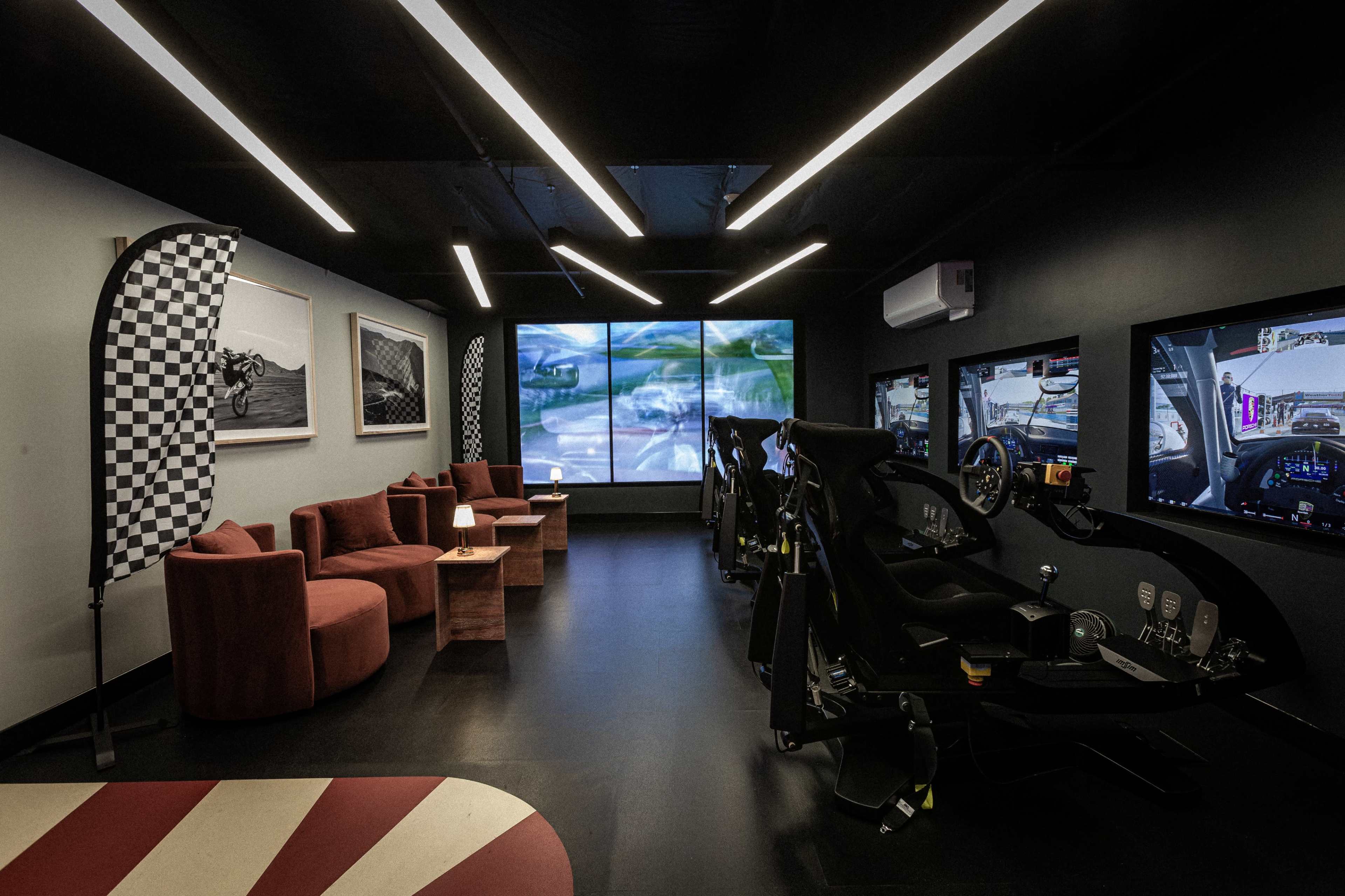 The image shows a sleek racing simulator room with two racing seats, large screens displaying a racing game, and cozy seating for spectators.
