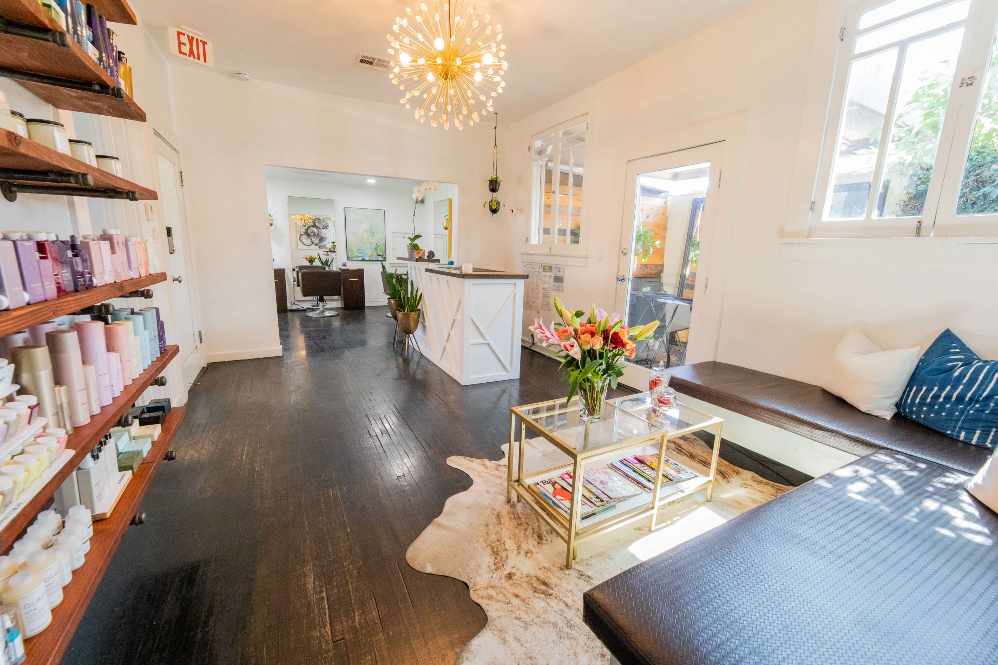 A bright, modern salon interior with a seating area featuring a glass coffee table, a large decorative light fixture, and shelves stocked with beauty products.