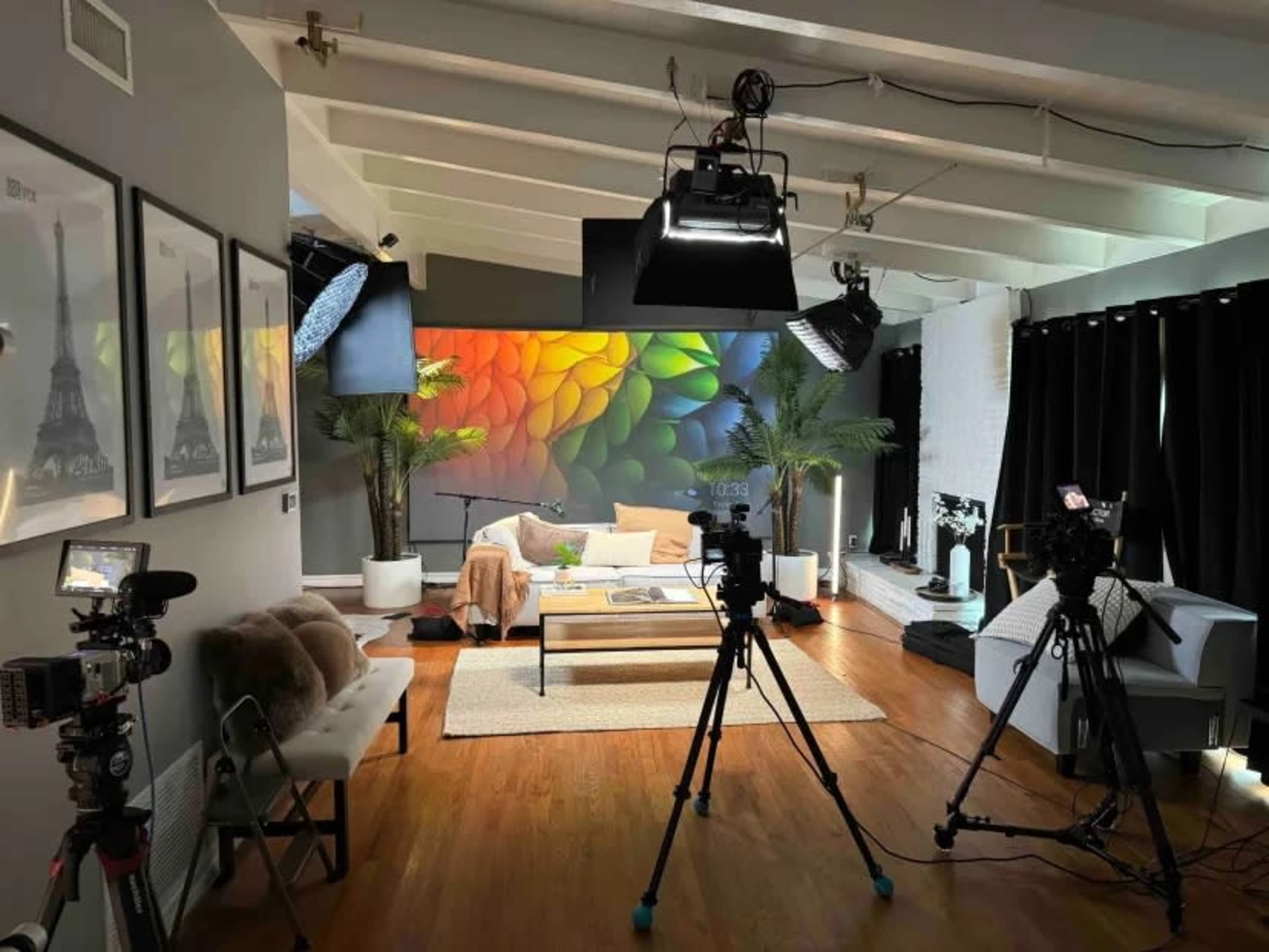 The image shows a well-lit indoor studio setup with cameras positioned around a modern living space featuring a colorful mural, plants, and a contemporary furniture arrangement.