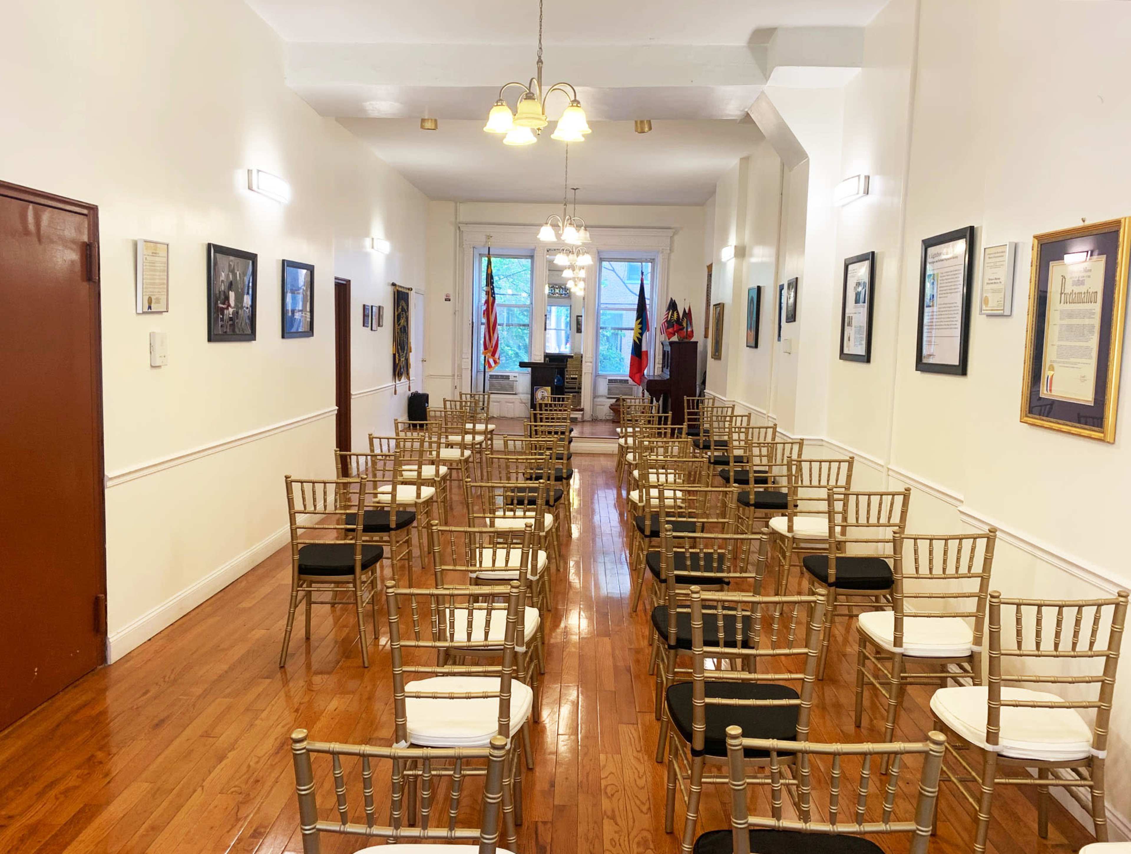 A long, narrow room with rows of gold chiavari chairs facing a front area, adorned with framed documents and flags along the walls.