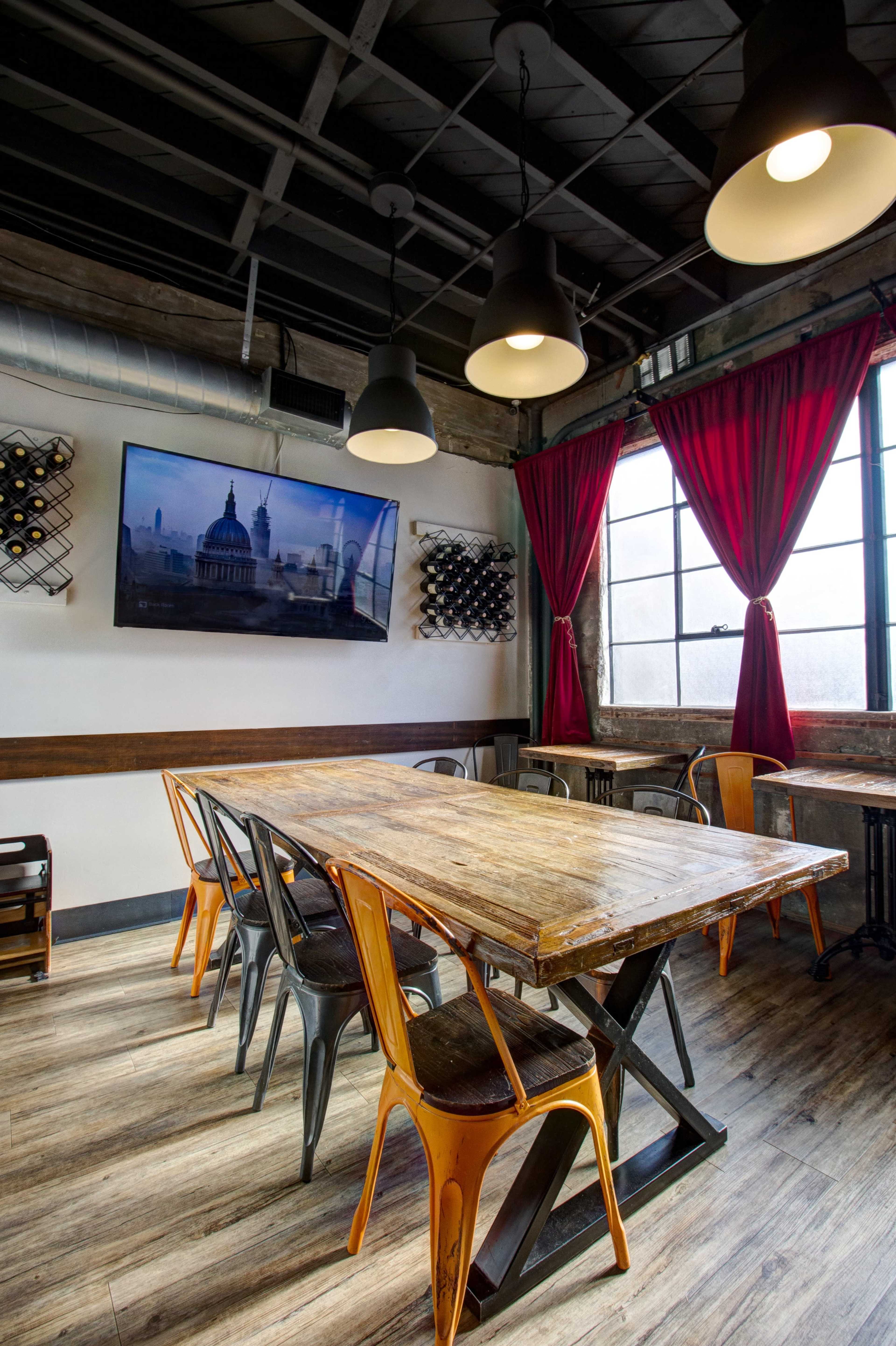 A dining area features a wooden table surrounded by metal chairs, with large windows draped in red curtains and a wall-mounted television displaying a cityscape.