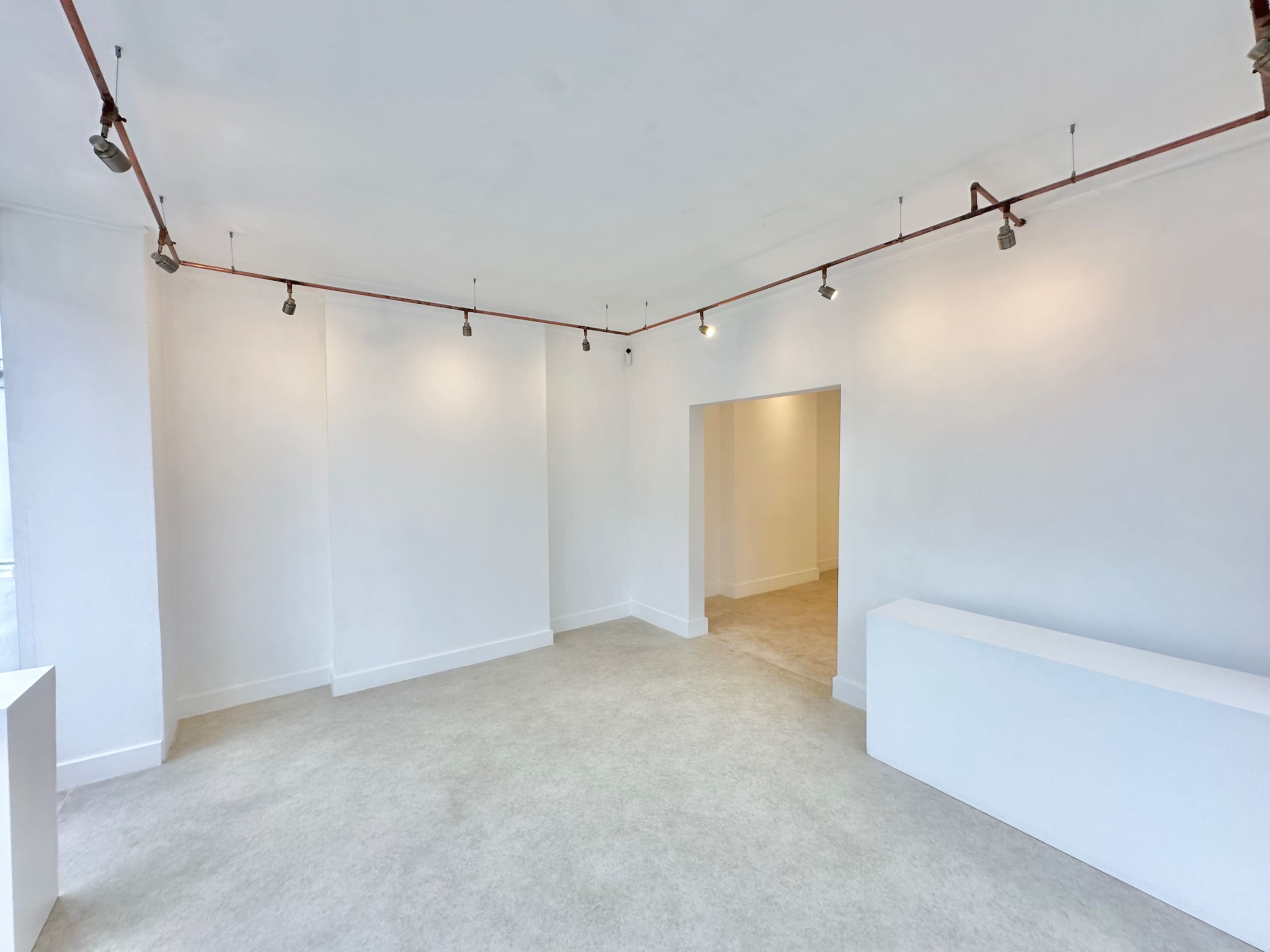The image shows an empty interior space with white walls, a light-colored floor, and exposed piping with track lighting.