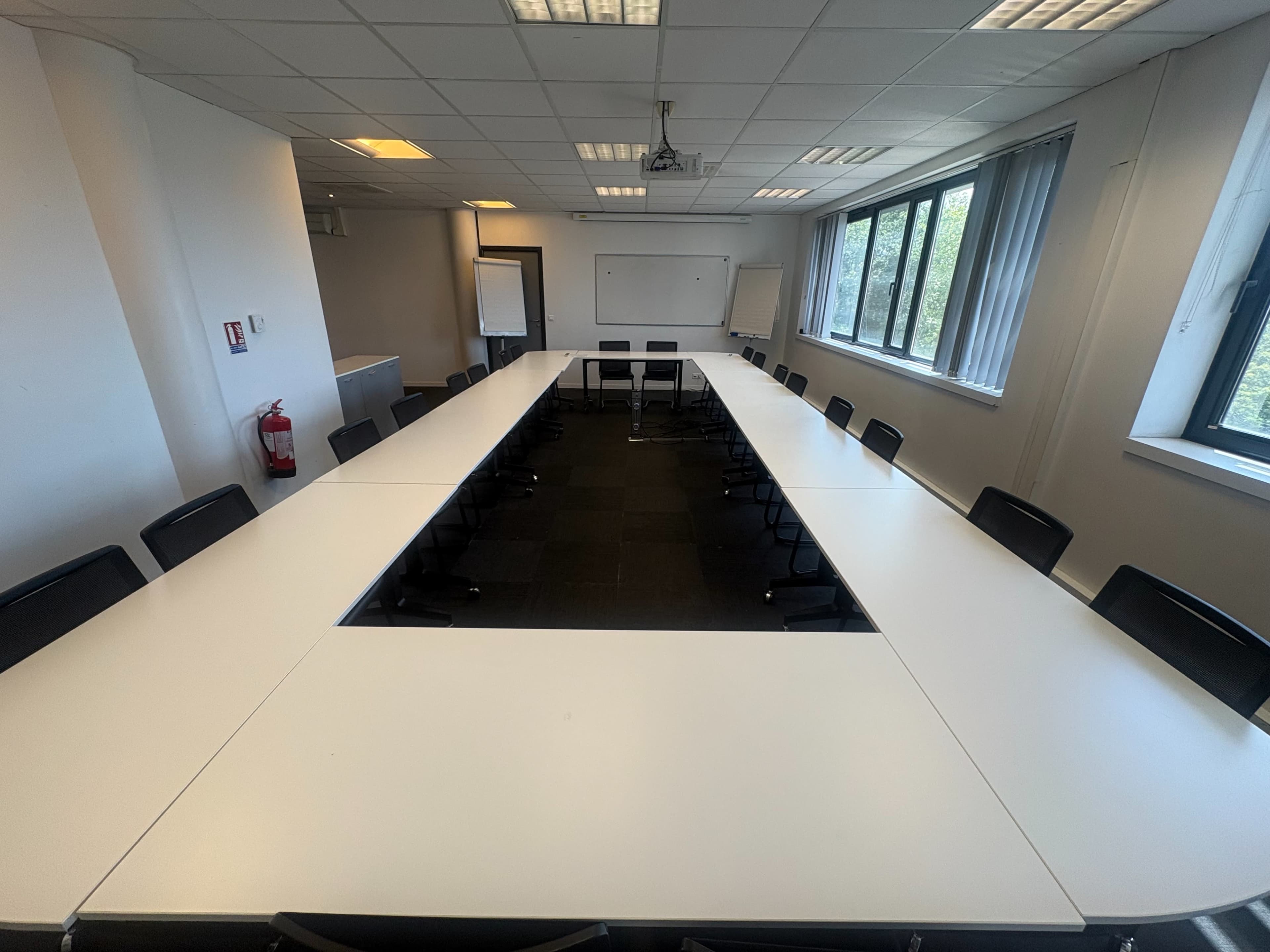 A large conference room features a rectangular arrangement of white tables surrounded by black chairs, with windows providing natural light.