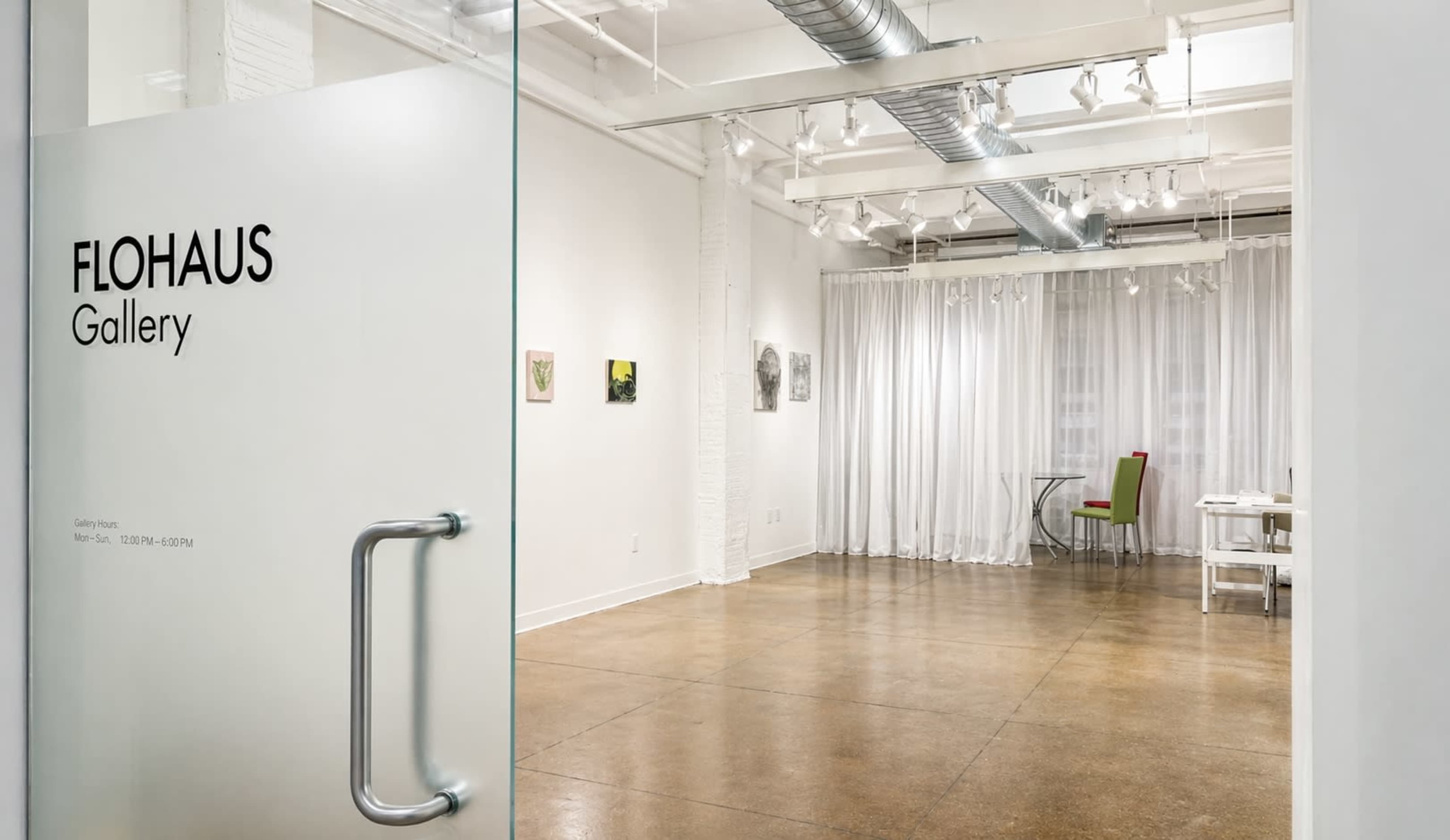 The image shows the interior of the Flohaus Gallery, featuring a bright, open space with white walls, artwork displayed on the walls, and a table in the distance.