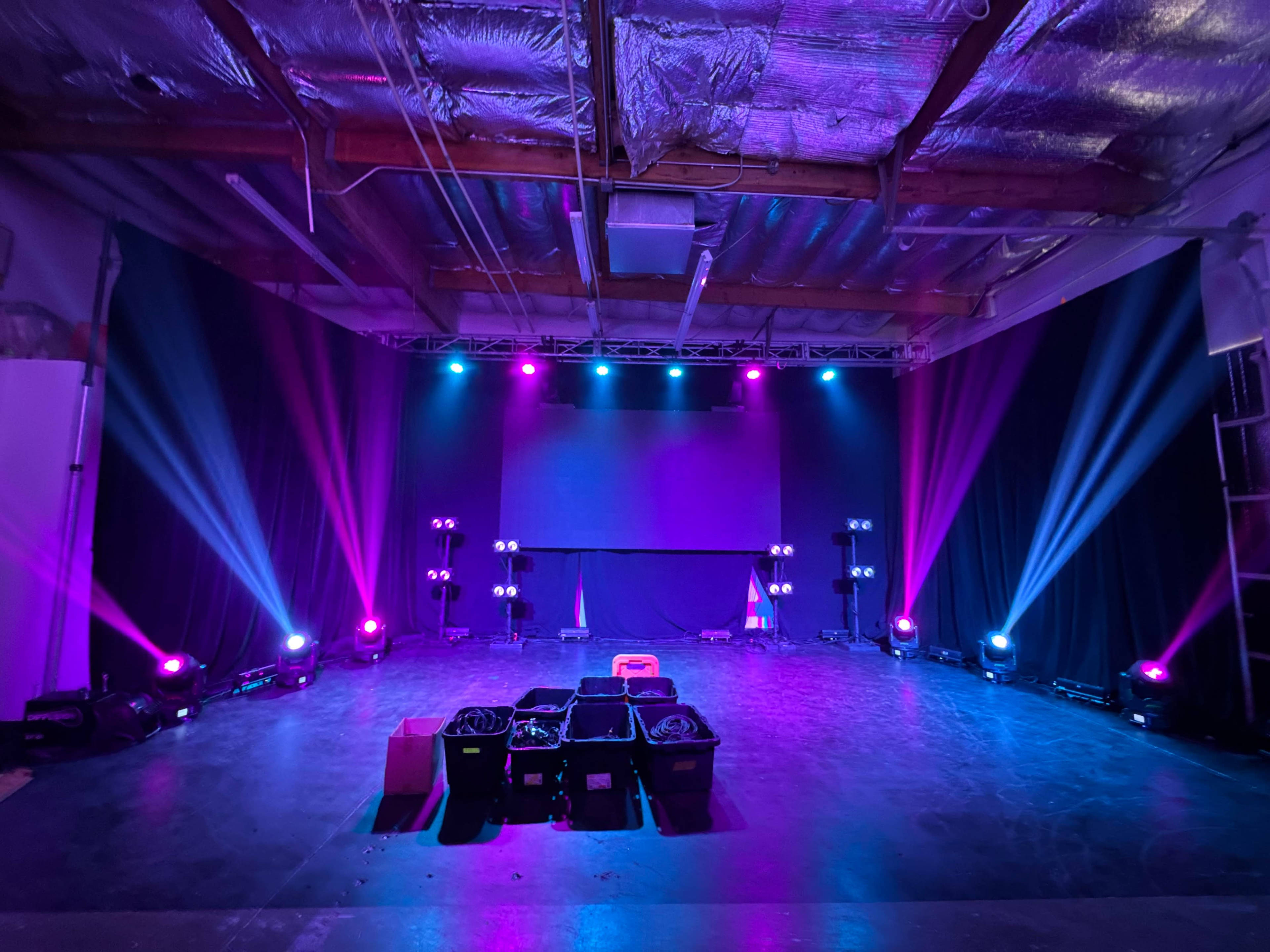 The scene features an empty stage with colorful lighting, a large screen in the background, and equipment stored in black bins on the floor.
