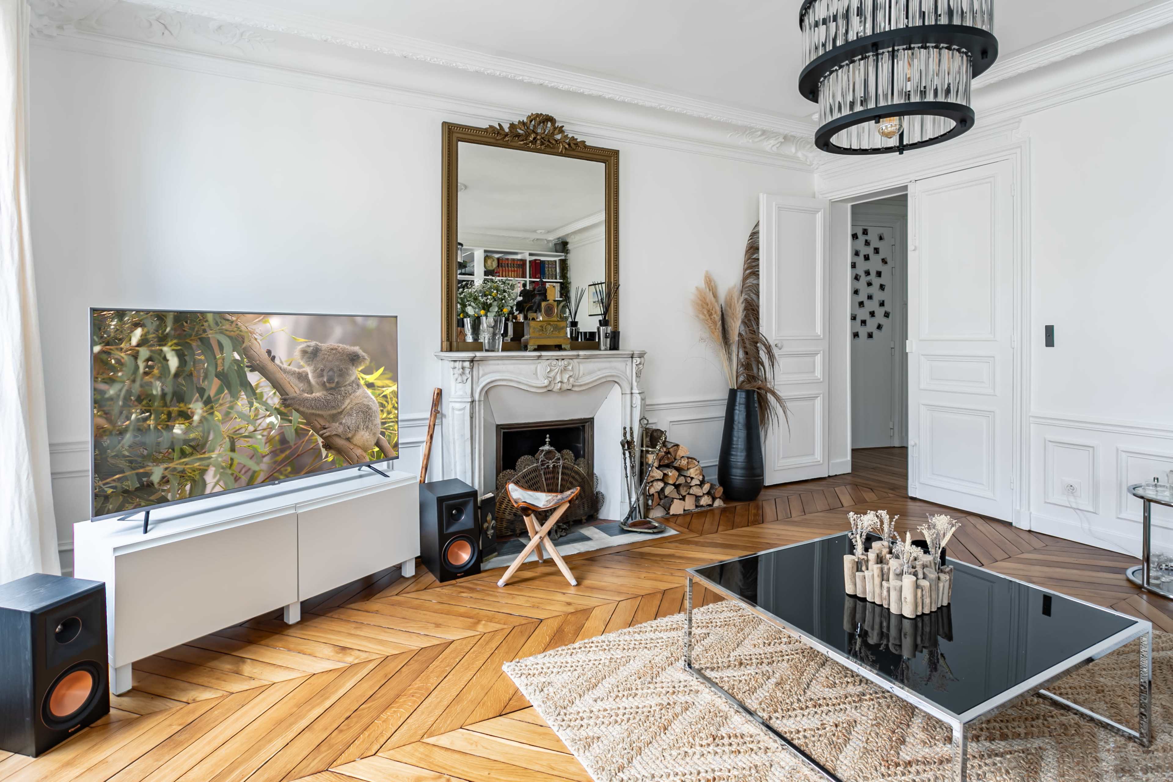 A modern living room features a television displaying a koala, a mirrored mantel above an elegant fireplace, and a coffee table surrounded by decorative elements.