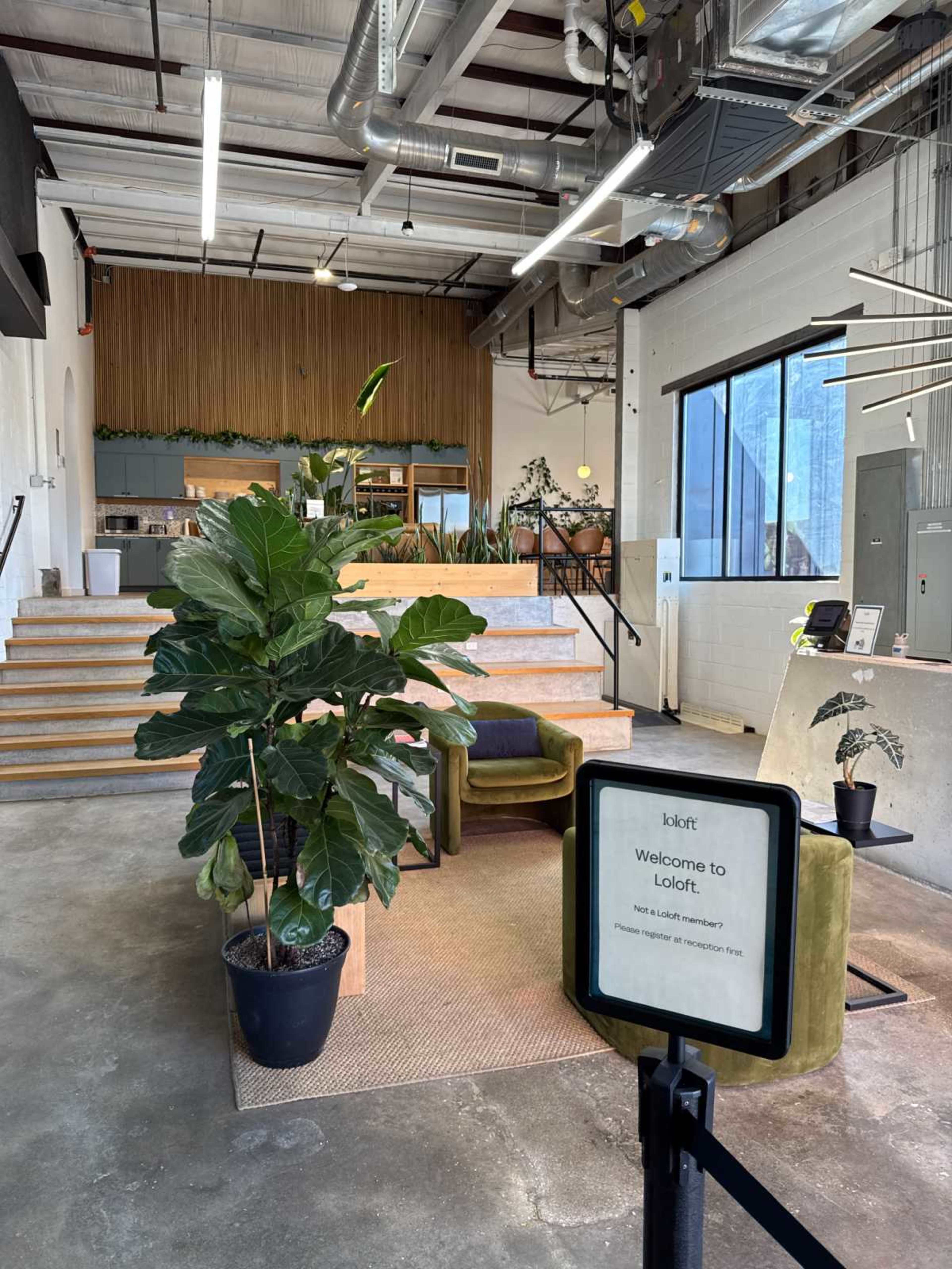 The image shows a modern lobby area with a reception desk, wooden accents, greenery, and seating on a concrete floor.