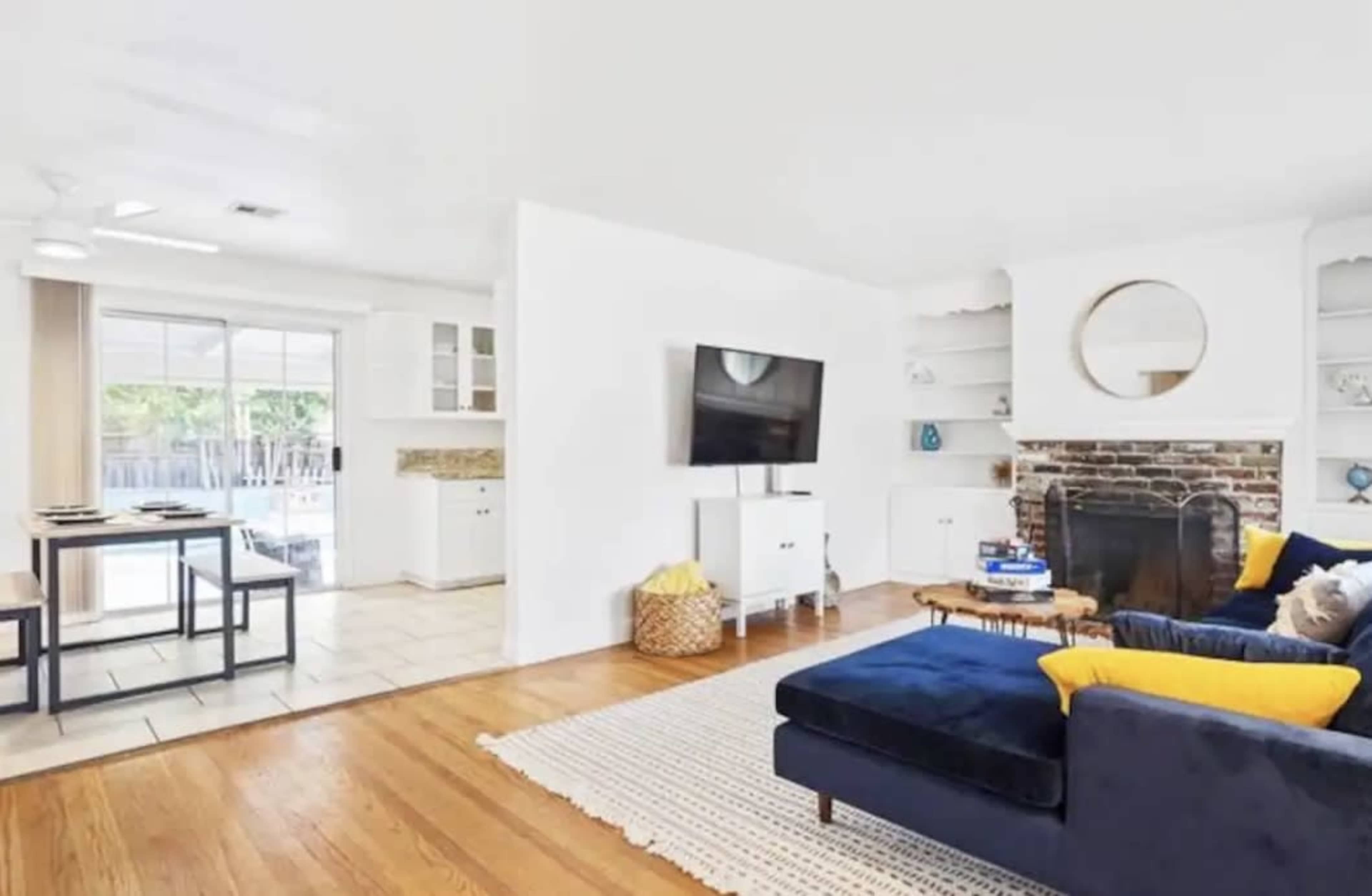 A living room features a dark blue sofa with yellow cushions, a television mounted on a white wall, and a brick fireplace surrounded by built-in shelves.