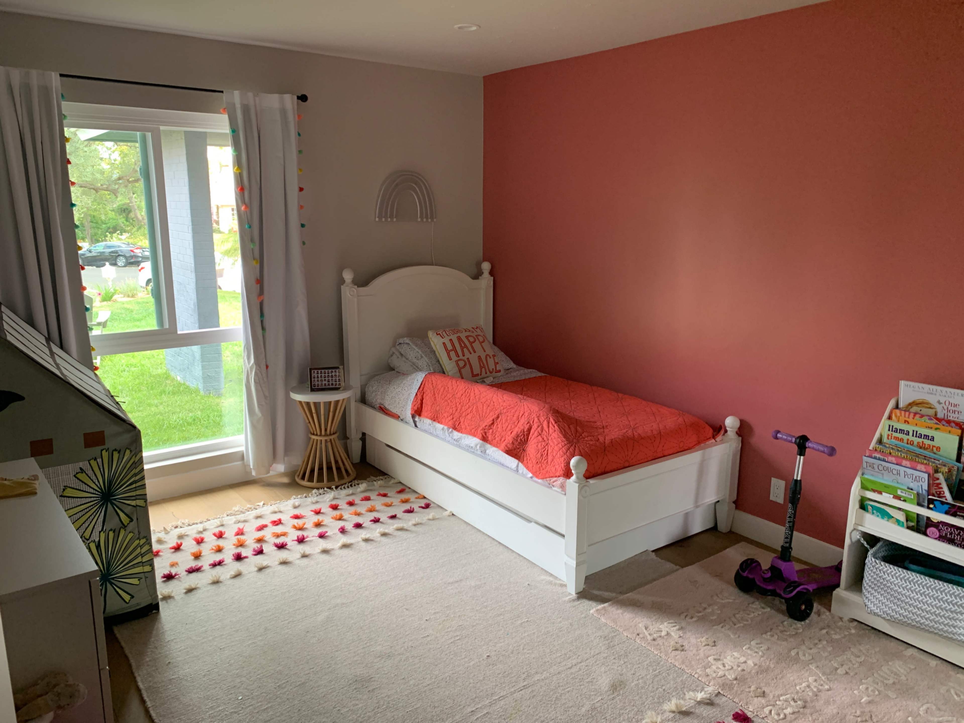 A children's bedroom featuring a white bed with an orange blanket, a small bedside table, and a scooter beside a colorful play area.