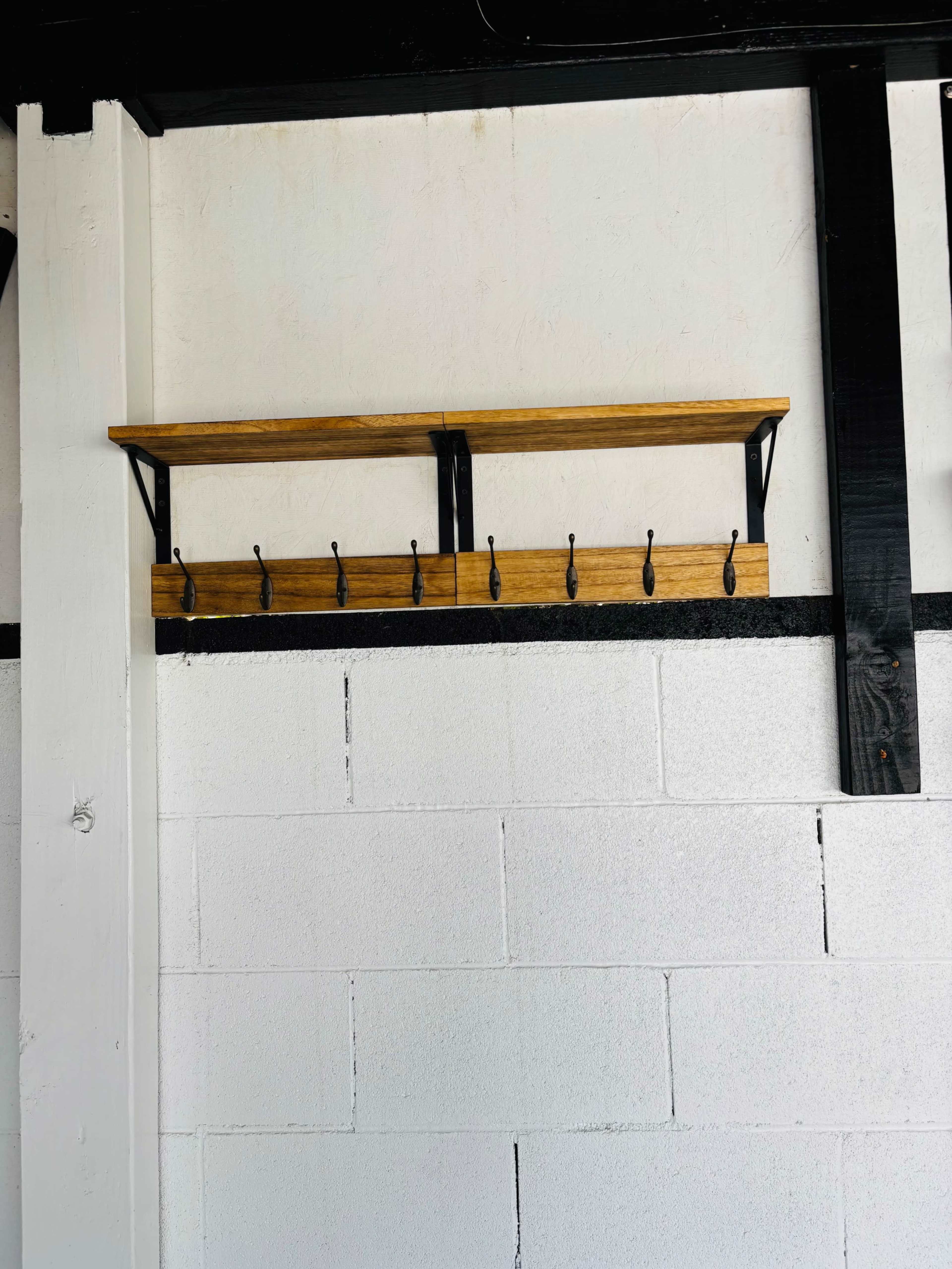 The image shows a wooden shelf with black metal brackets mounted on a white cinder block wall, featuring several hooks underneath.
