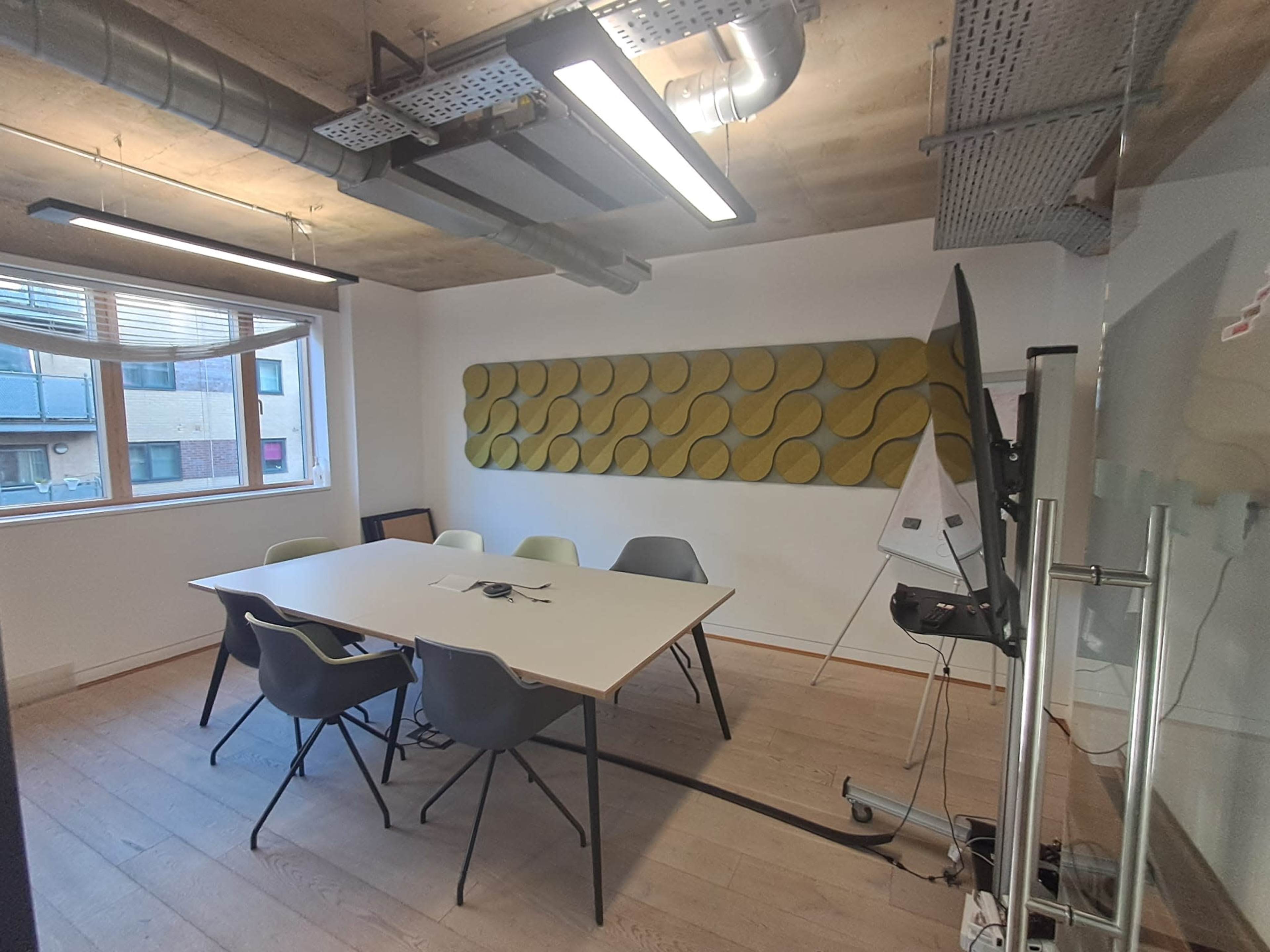 The image shows a modern conference room with a long table surrounded by chairs, a large window, and a textured wall panel.