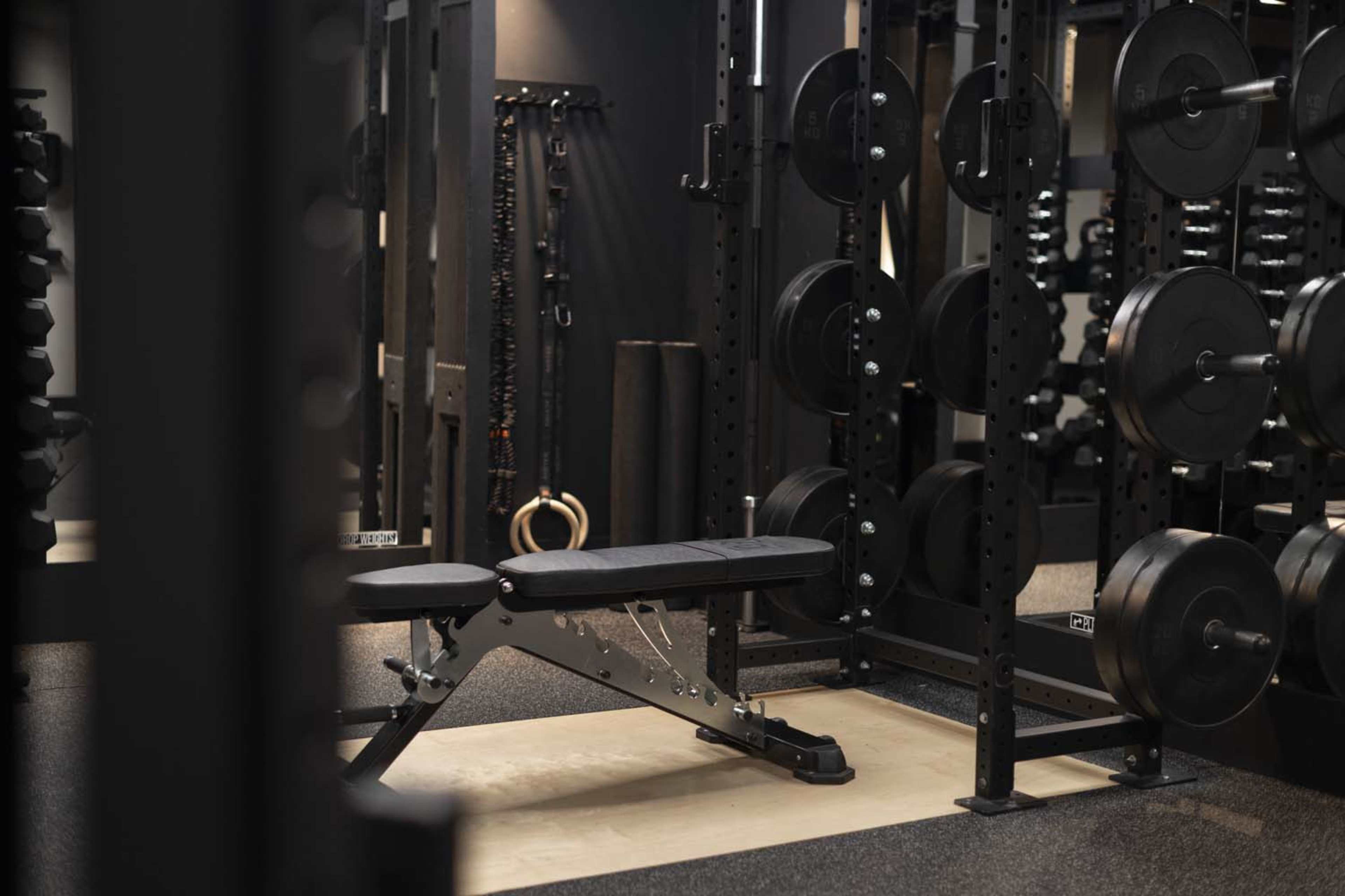 The image shows a well-equipped gym with a weight bench positioned in front of a rack filled with weight plates.
