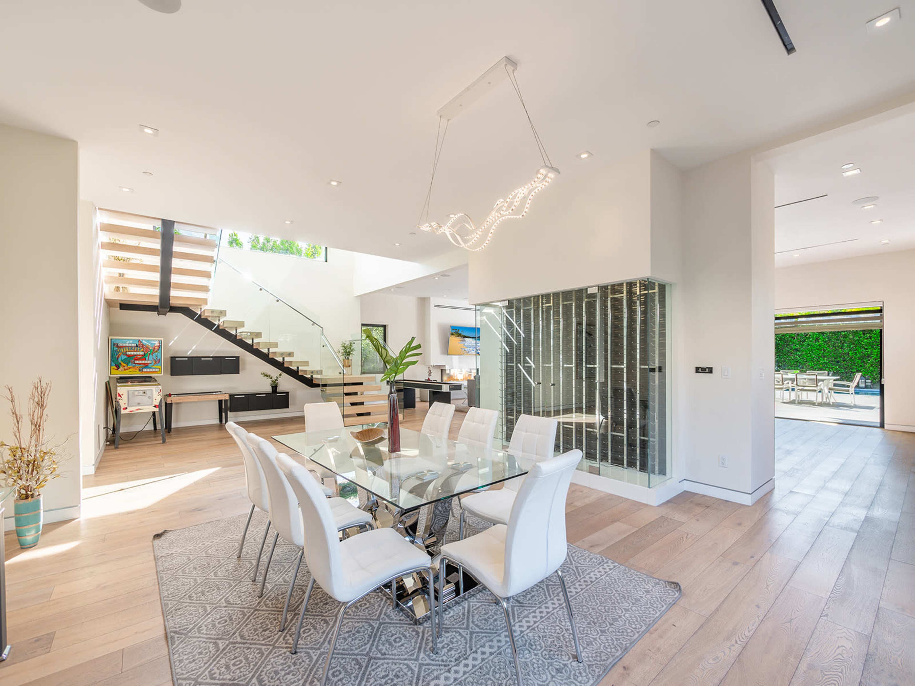 A modern dining area features a glass table surrounded by white chairs, with a staircase and large windows allowing natural light to illuminate the space.