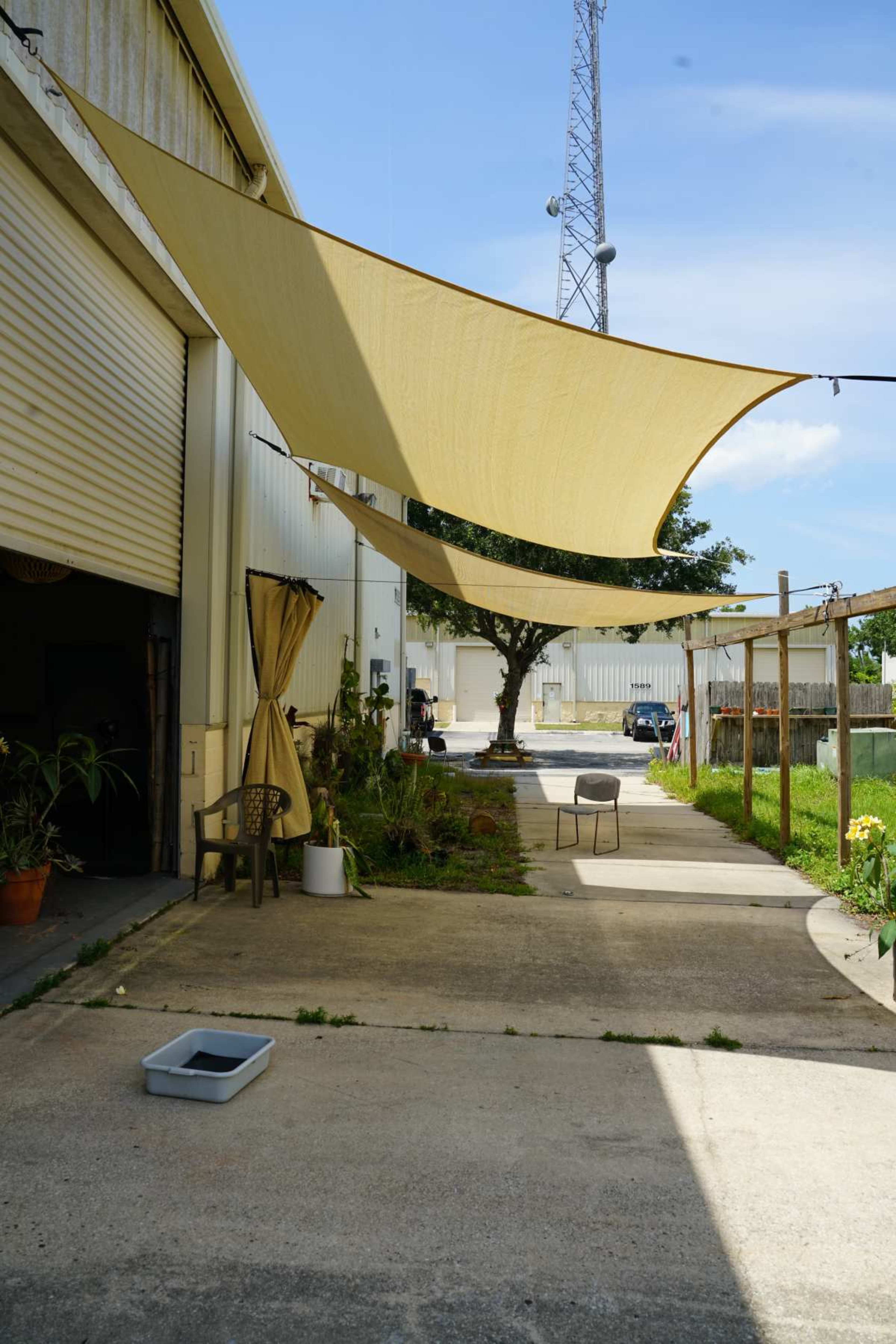 The image shows a narrow walkway between buildings, covered by large fabric shades, with pots, a chair, and a tree in the foreground.