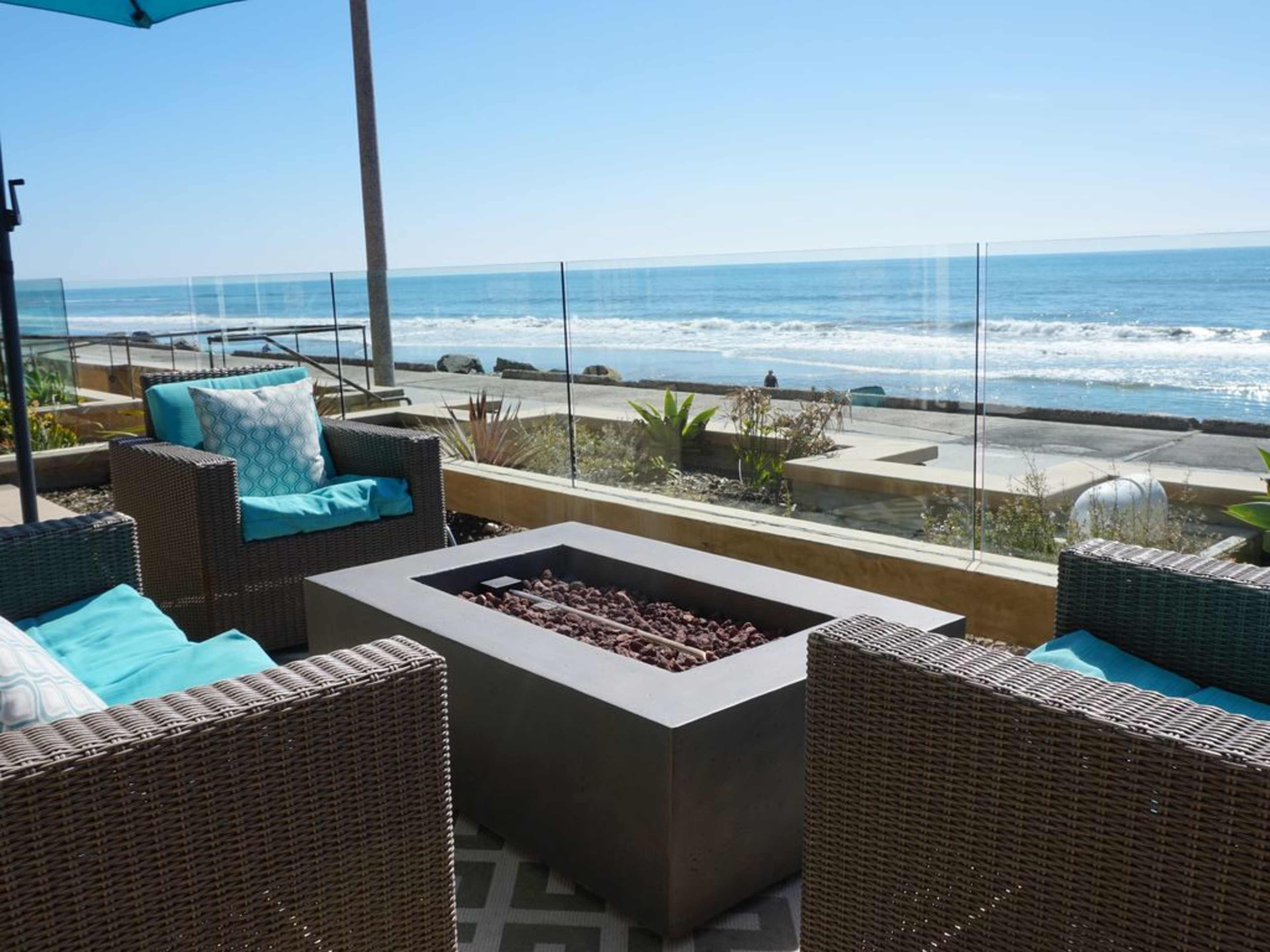 A modern outdoor seating area features two wicker chairs with blue cushions, surrounding a rectangular fire pit, overlooking the ocean.
