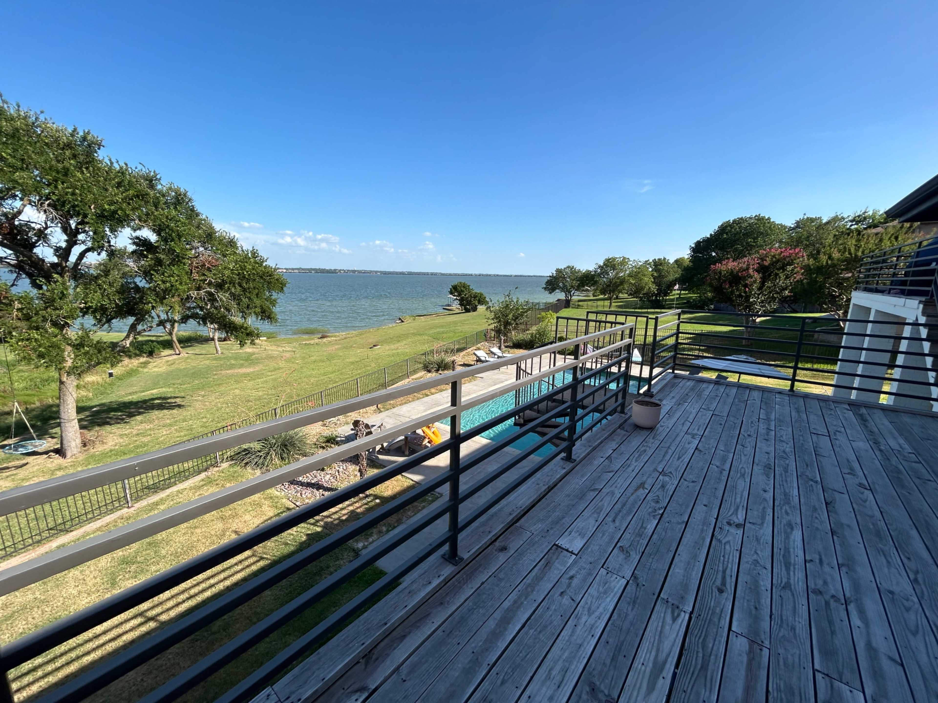 A wooden deck overlooks a grassy area and a body of water, with trees and a swimming pool visible in the background.