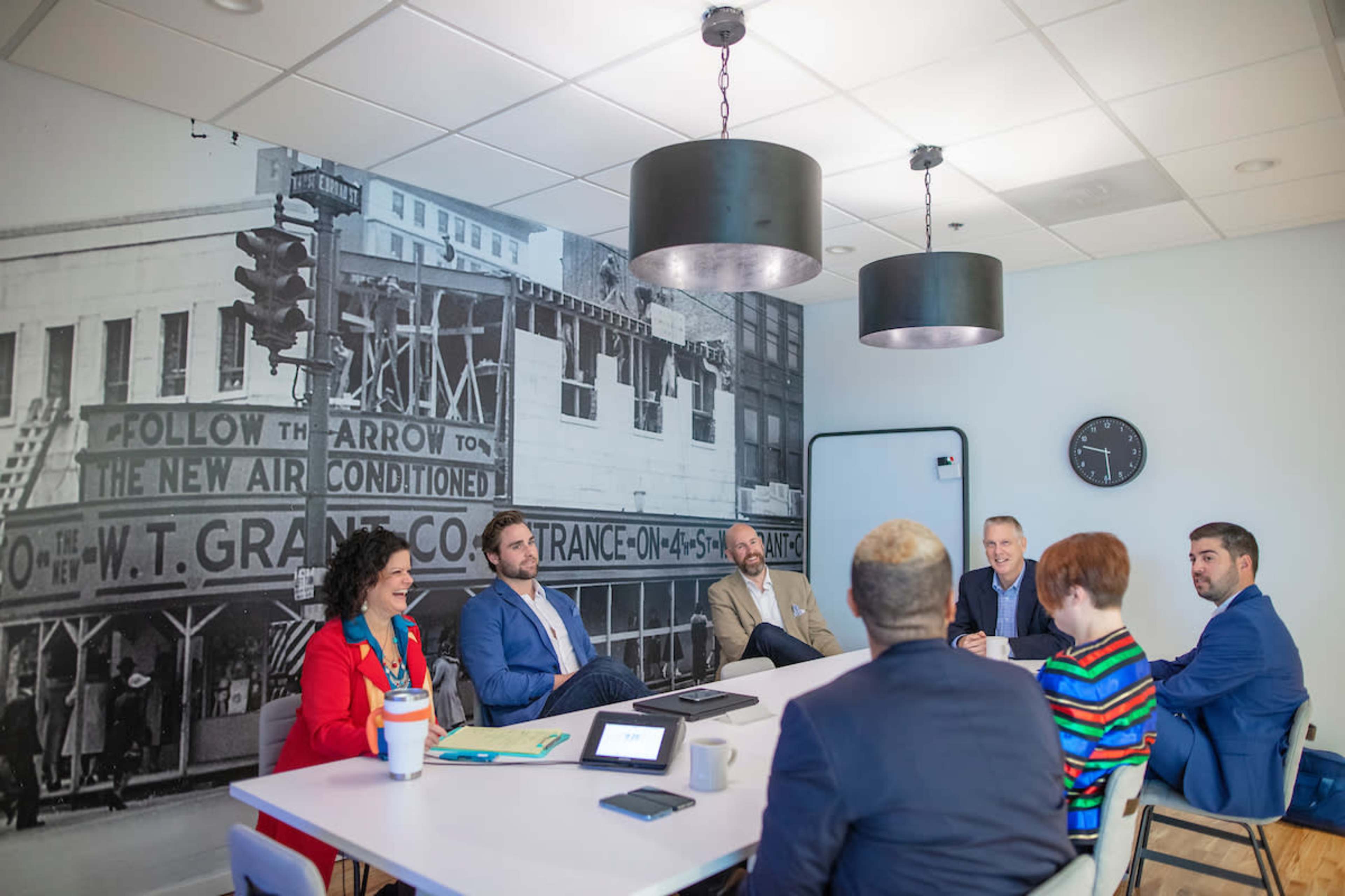 A diverse group of professionals is engaged in a discussion around a conference table in a room featuring a large historical black-and-white mural.