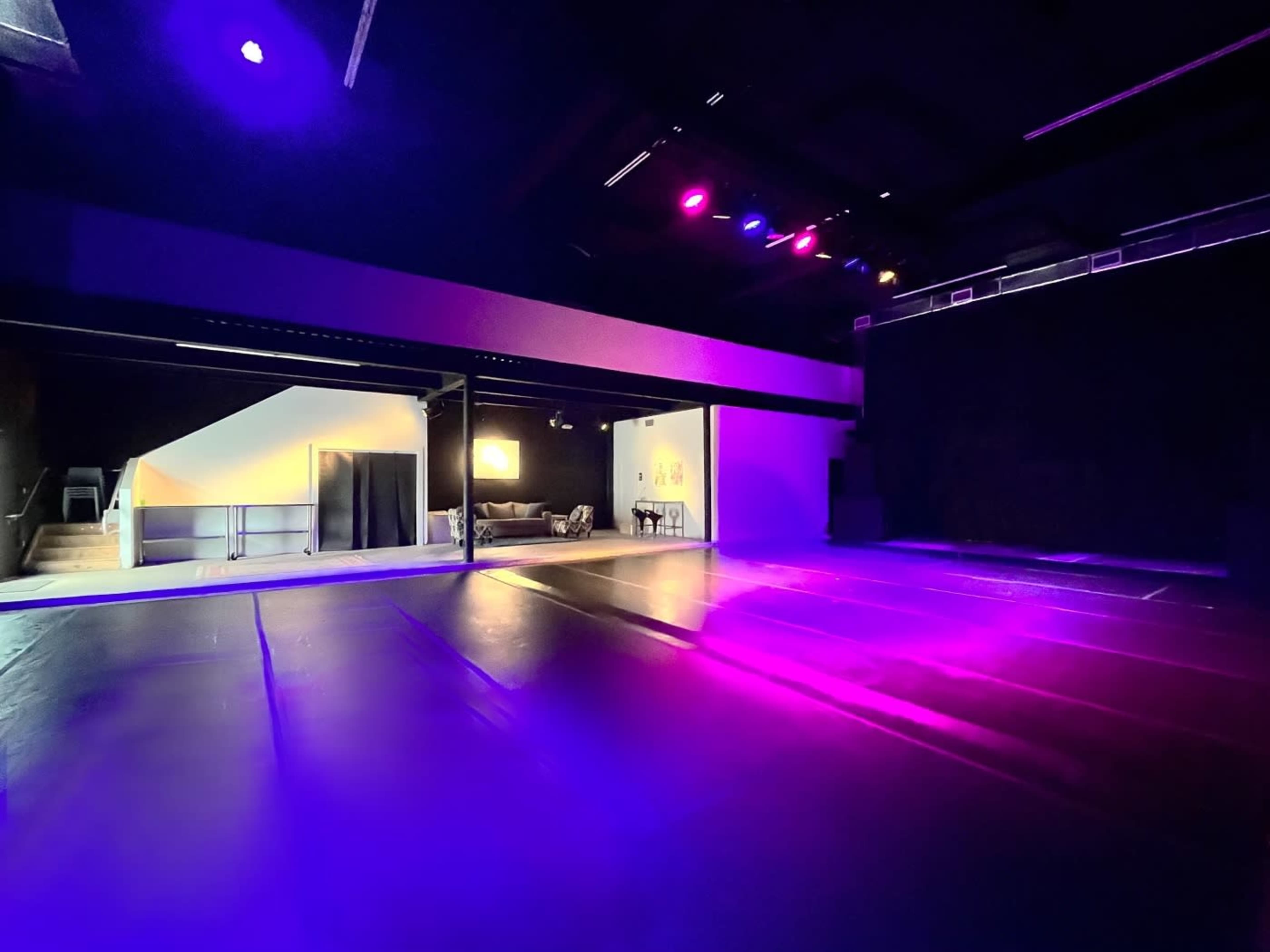 The image shows an indoor performance space with dim lighting, featuring a stage area with purple lights and a nearby room that appears to have seating and decor.