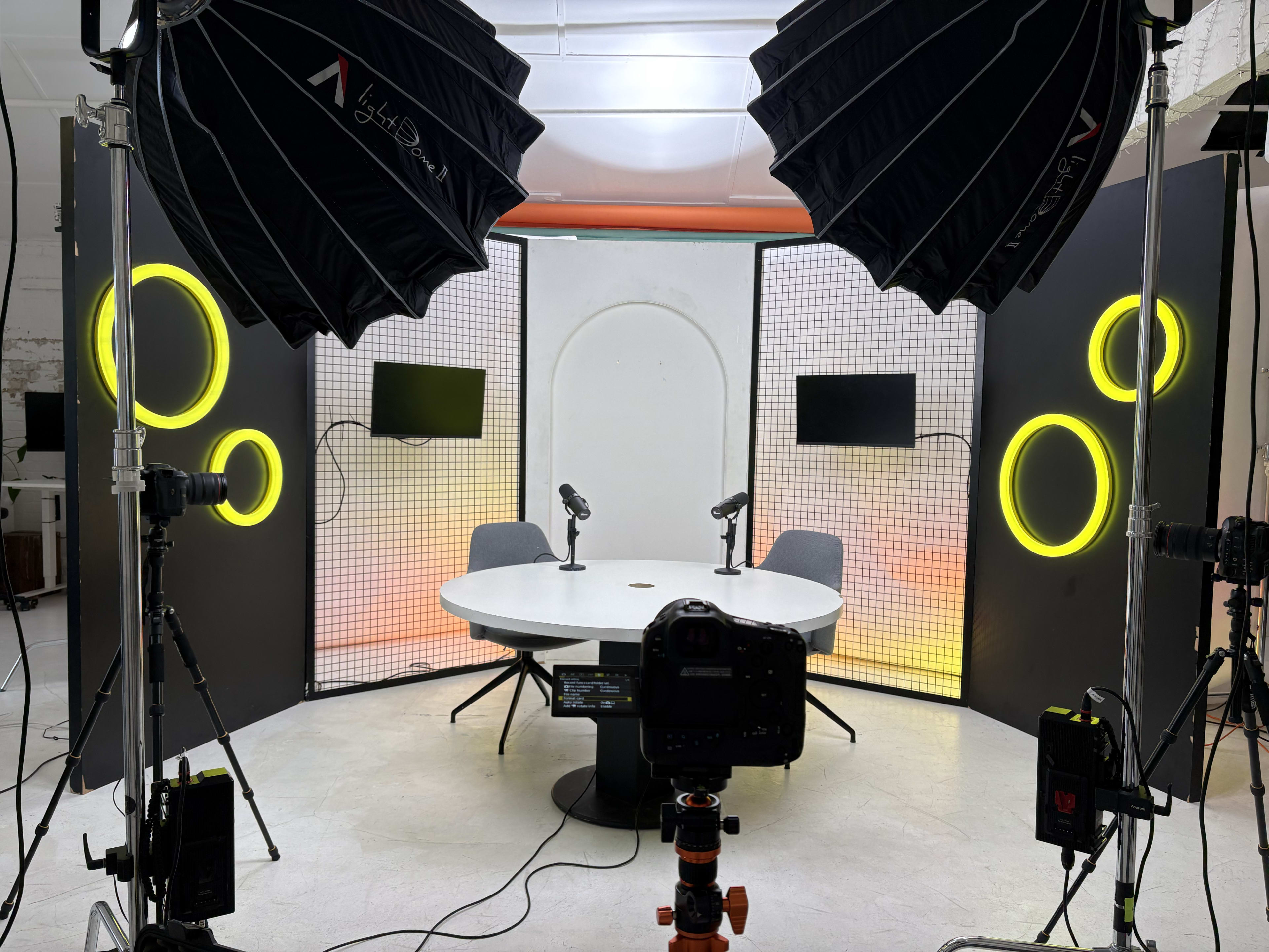 The image shows a modern studio setup with a circular table, two microphones, illuminated backdrops featuring large yellow circles, and multiple cameras and lighting equipment.