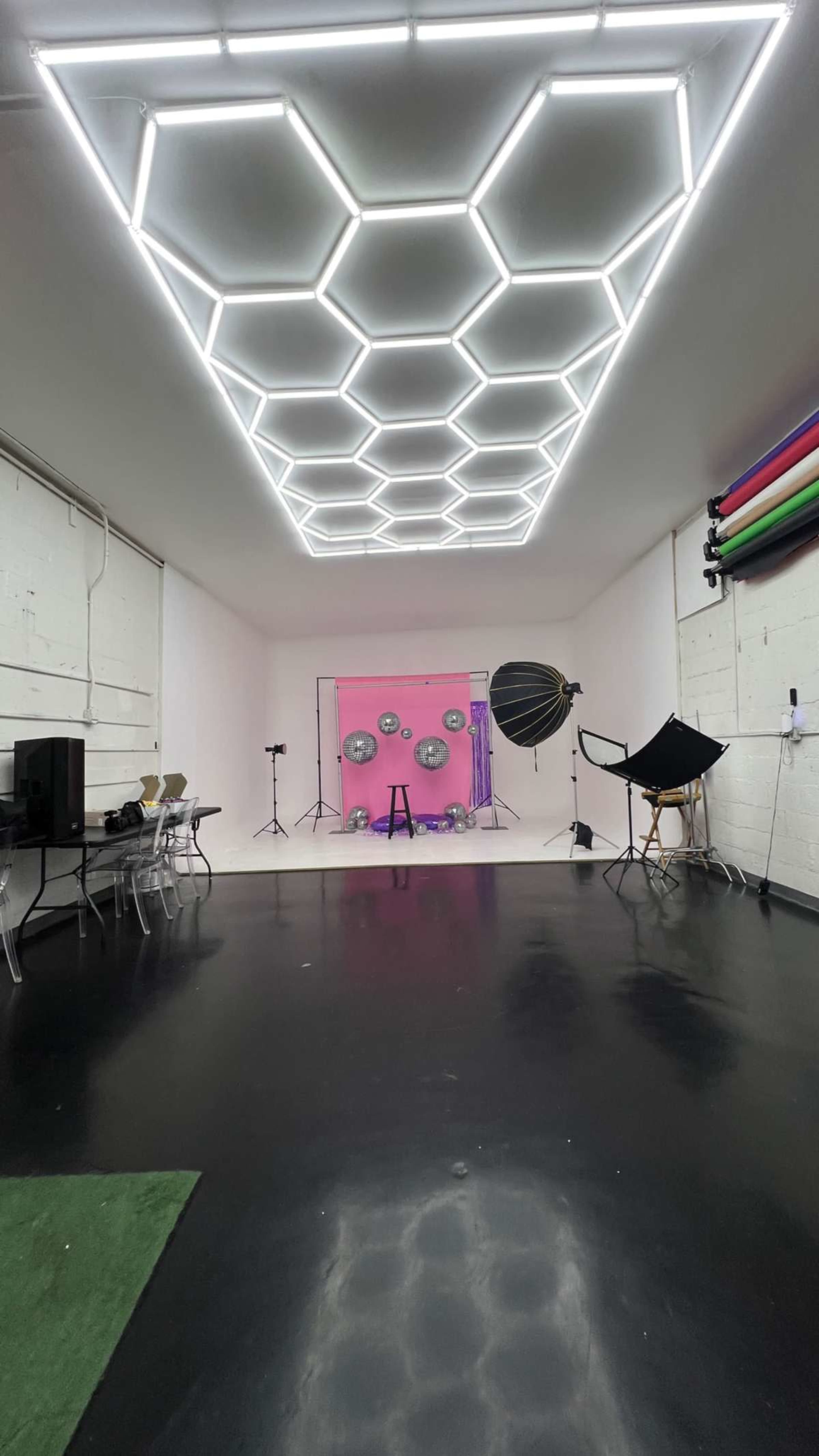 The image shows a spacious studio with a hexagonal light ceiling, black flooring, a pink backdrop, and various photography equipment, including a large umbrella light and a chair.