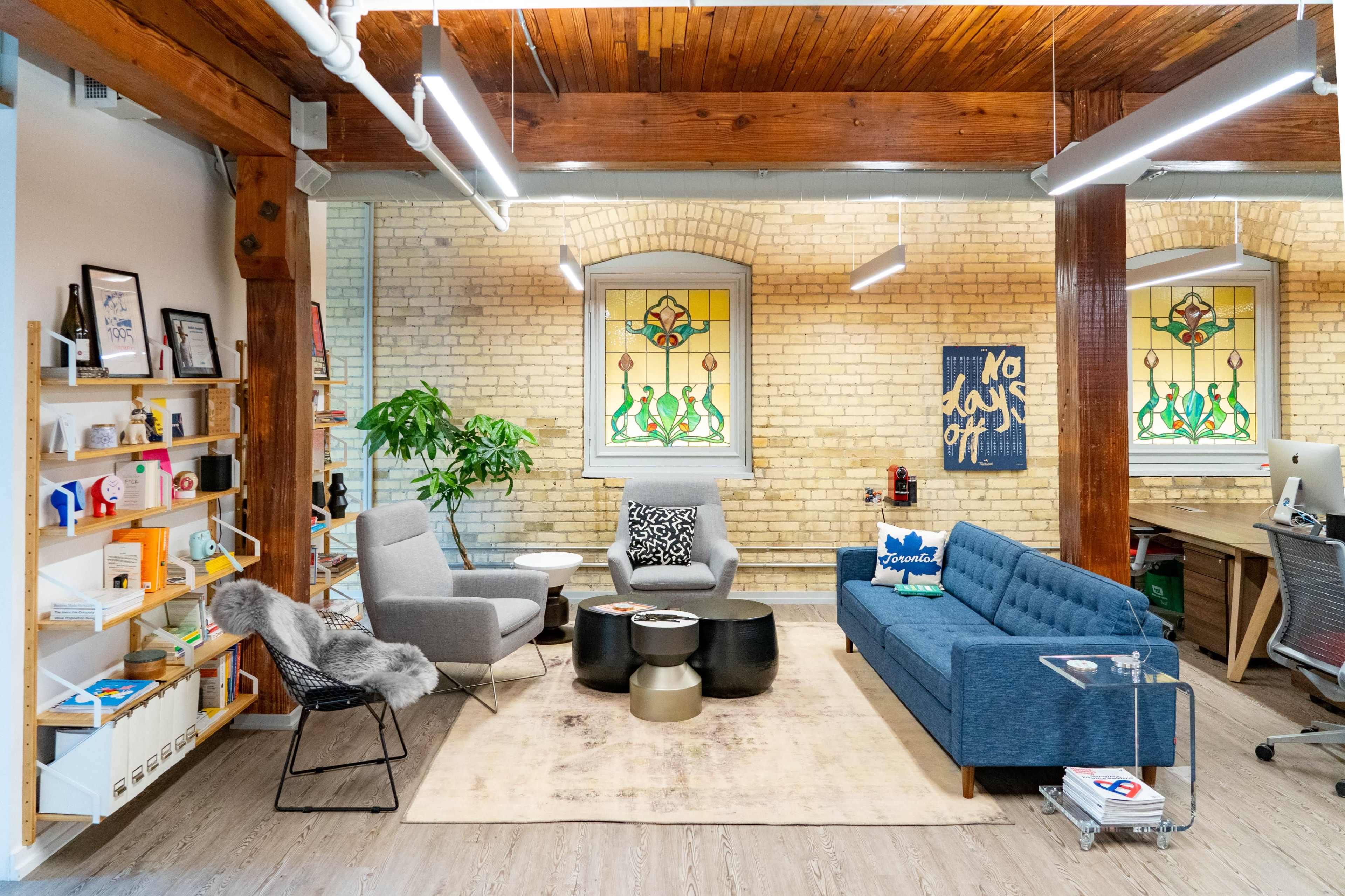 A modern office space featuring a seating area with a blue couch, gray chairs, a coffee table, and large windows with decorative stained glass.